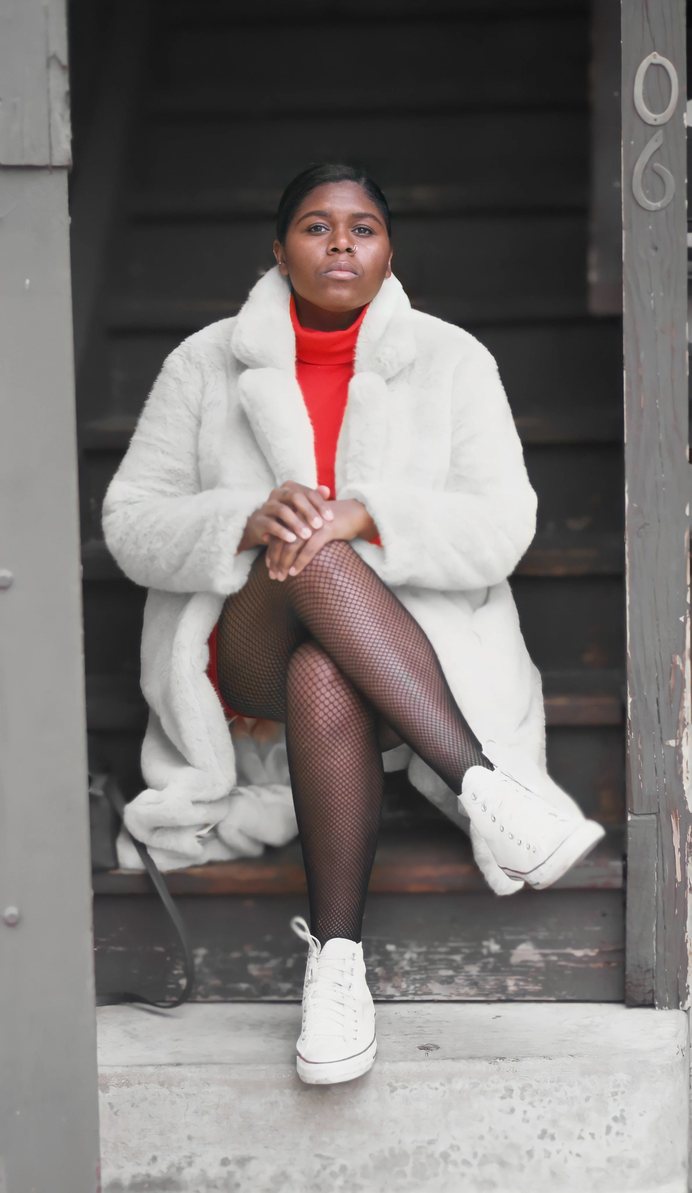 Woman sitting on wooden steps, wearing a white faux fur coat, red turtleneck, fishnet stockings, and white sneakers, with crossed legs and hands resting on knee