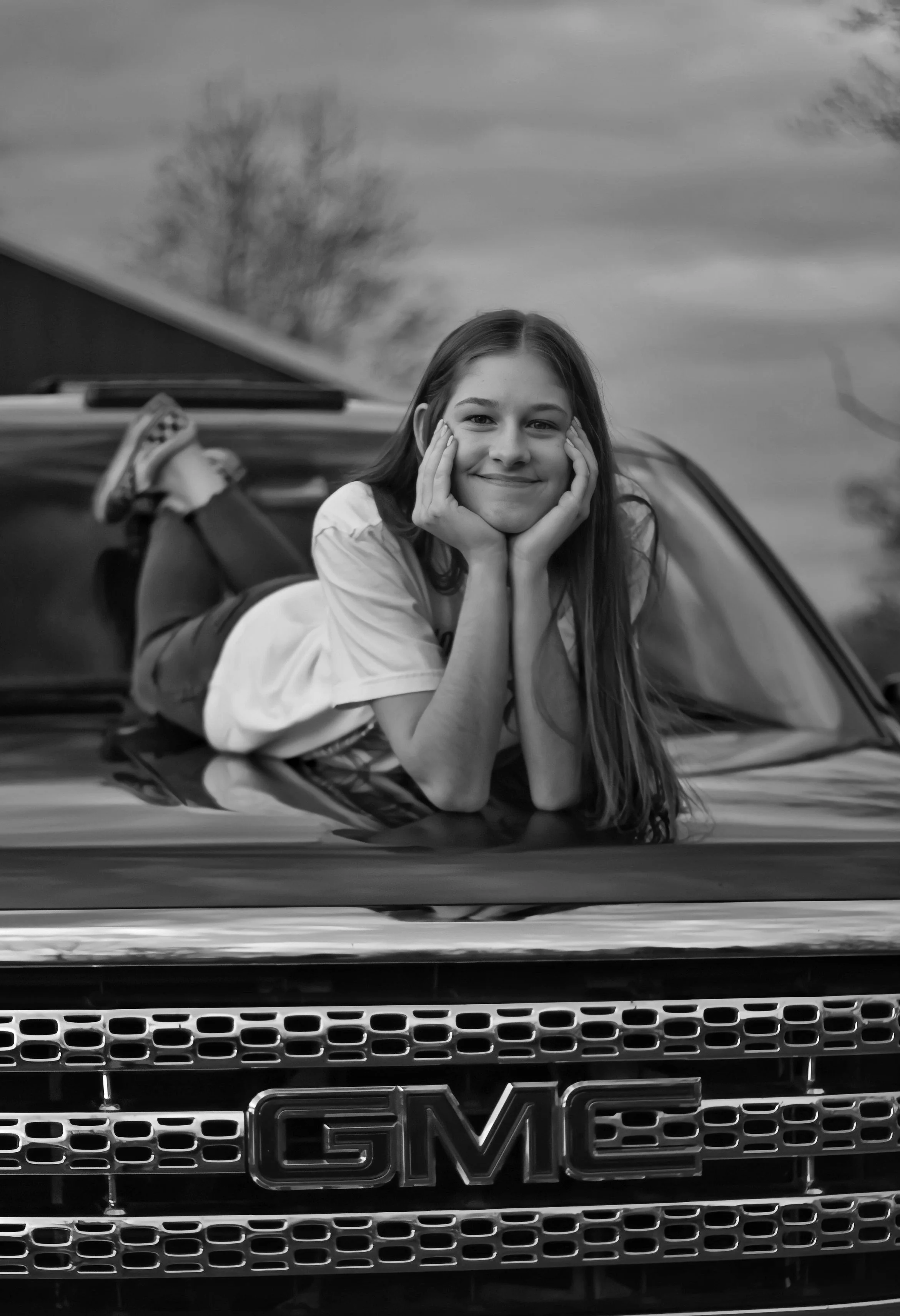 A young woman with long hair lying on the hood of a GMC vehicle, resting her chin on her hands and smiling, with trees and cloudy sky in the background.