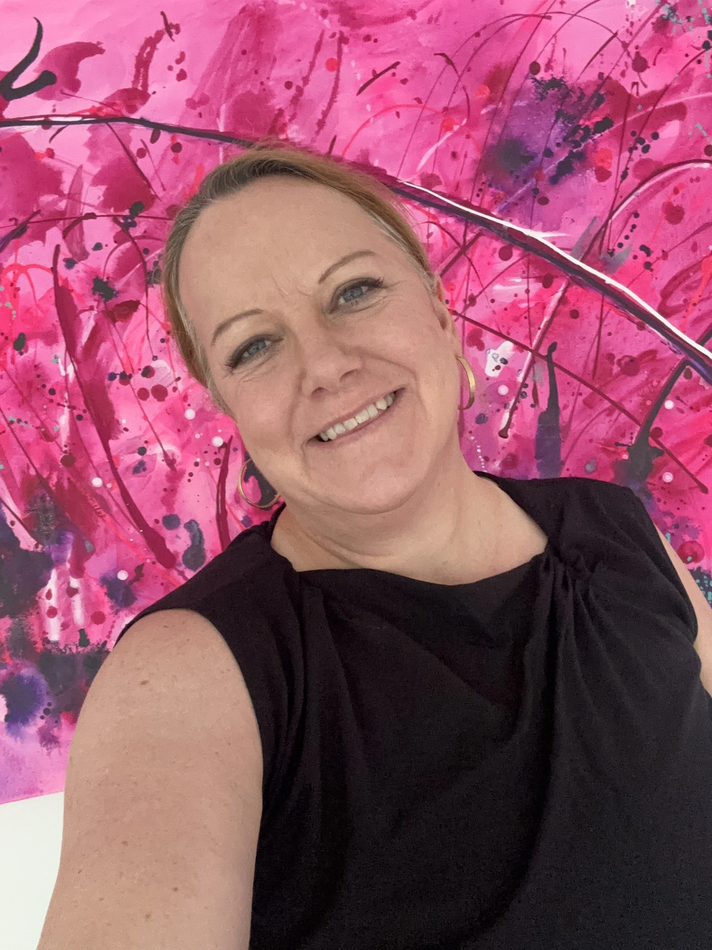 A woman smiling in front of a vibrant pink abstract painting with splashes of darker pink and black.