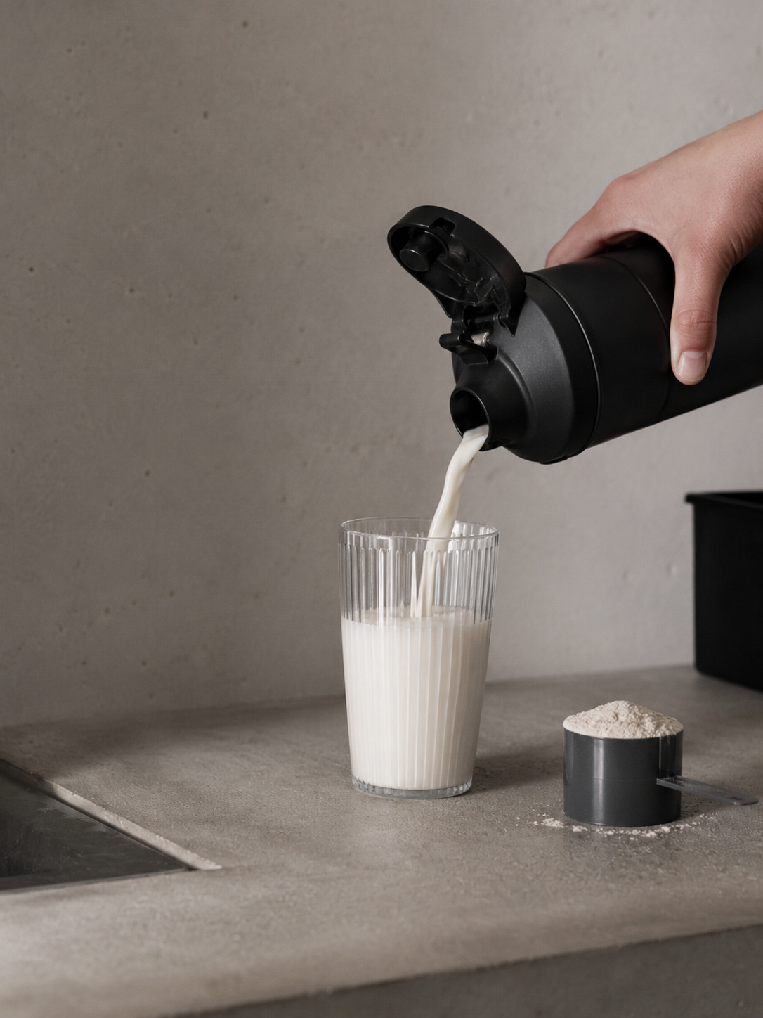 A hand pouring milk from a black electric milk frother into a clear, ribbed glass on a gray countertop. There is a scoop of white powder and a small measuring spoon nearby on the countertop.