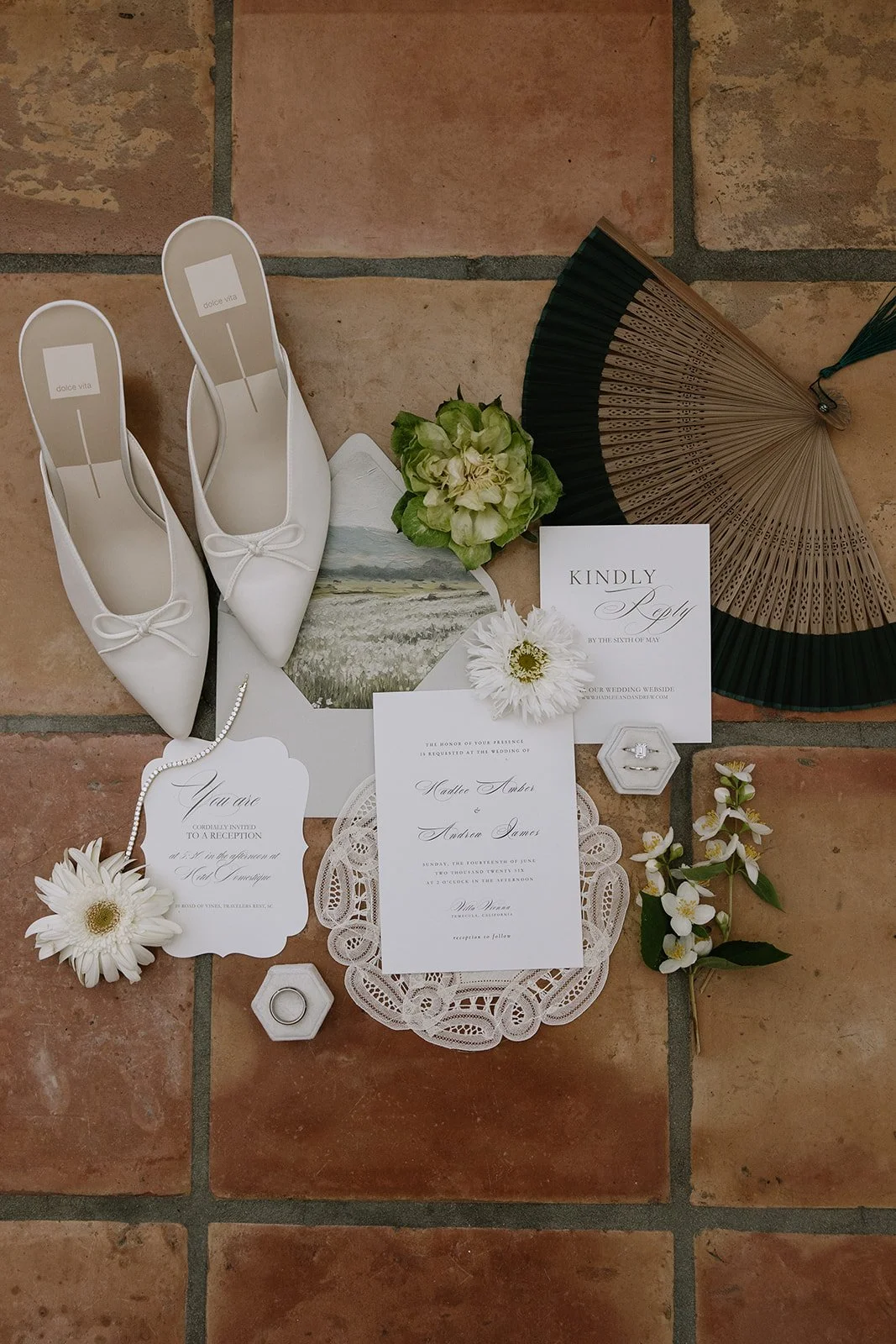 Wedding flat lay with white bridal shoes, green and white flowers, a black and gold hand fan, wedding invitations, a ring in a hexagon box, and decorative lace and paper accessories on a tile floor.