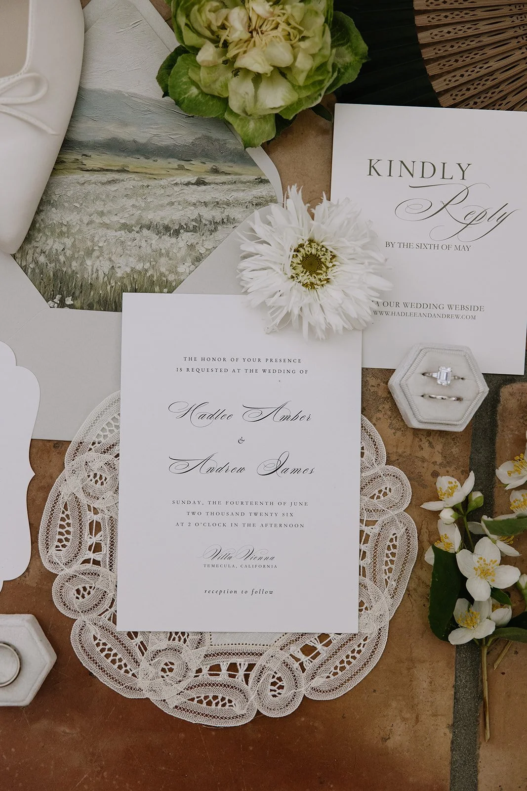 Wedding invitation suite with floral accents, includes an invitation card, an RSVP card, a details card, a white flower, lace doily, a small ring box with rings, and various flowers and greenery. Vintage inspired and romantic handwritten details.