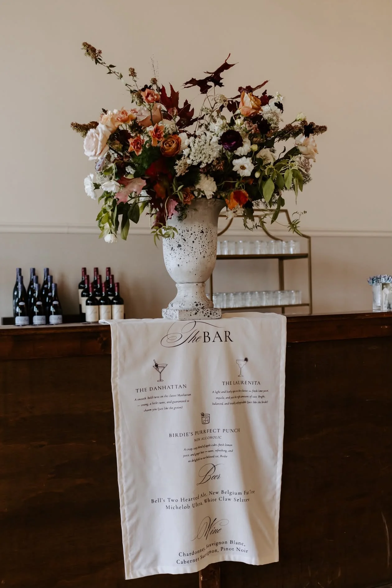 A fabric bar sign hanging from the bar reads 'The Bar' with the names and descriptions of cocktails and beers. Perfect for a wedding open bar to share the couples' signature drinks and drink menu.