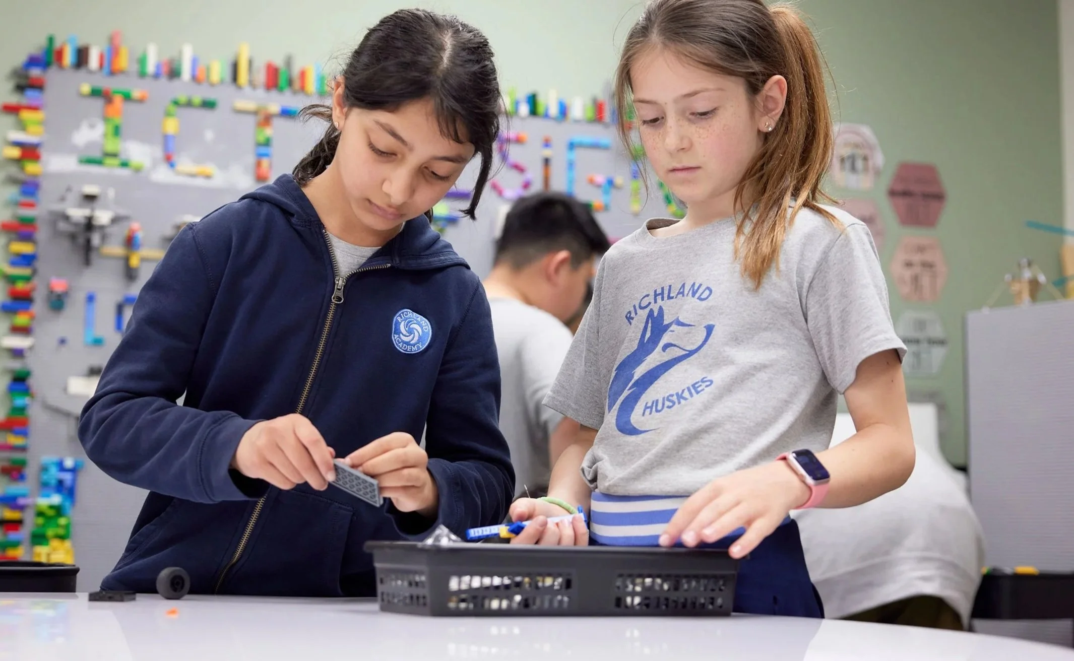 Two young girls working together on a robotics project in a classroom, with a colorful pegboard in the background.