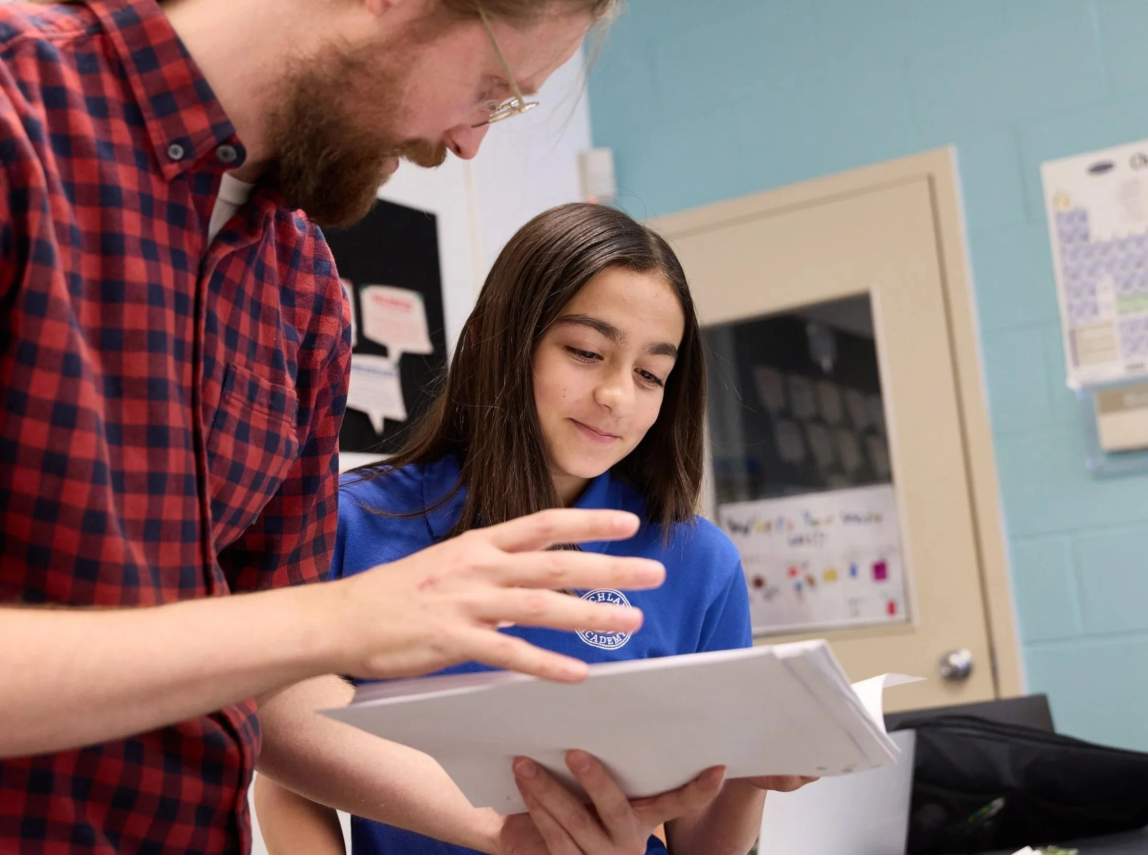 A young girl with long dark hair and a light tan complexion is reading a book or paper. An adult male with glasses, a beard, and fair skin is leaning over her, looking at the same paper. The background shows a school or classroom setting with posters on the wall.