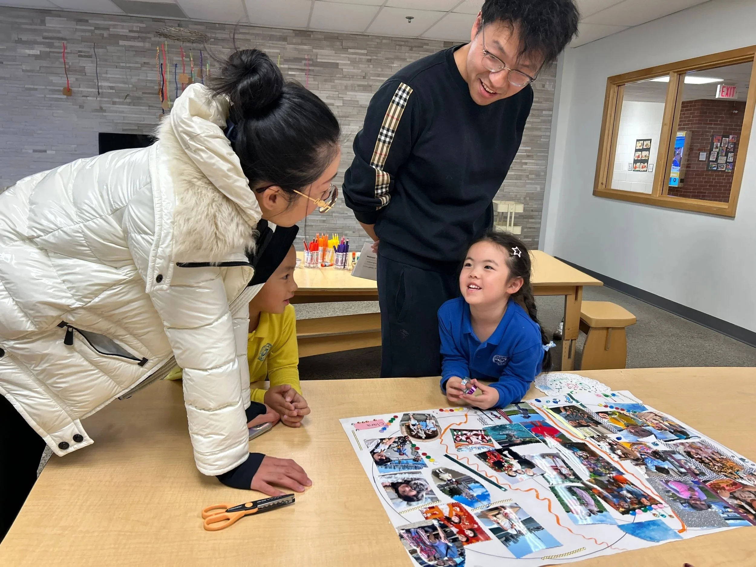 A group of people, including a young girl, an adult woman, and an adult man, are gathered around a table looking at colorful photo collage and craft supplies. The young girl is smiling while holding a small item, and the adults are engaged and smiling.