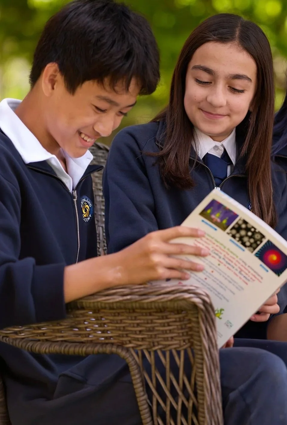 Two students in school uniforms sitting outdoors, smiling and looking at a pamphlet or brochure.