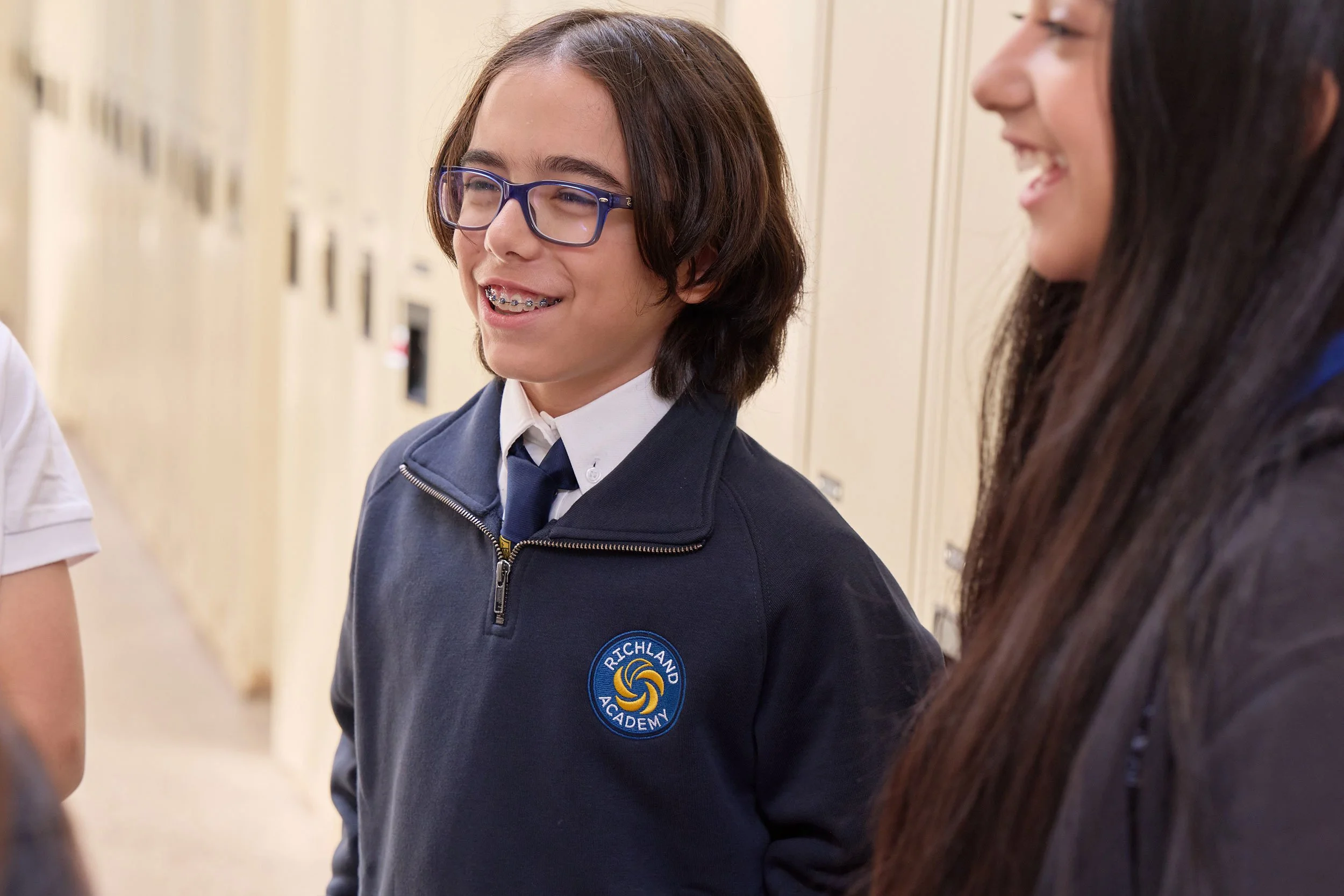 A young girl with shoulder-length brown hair, wearing glasses and a dark blue school uniform with a 'Richland Academy' badge, smiling and talking to another girl with long dark hair in a school hallway.