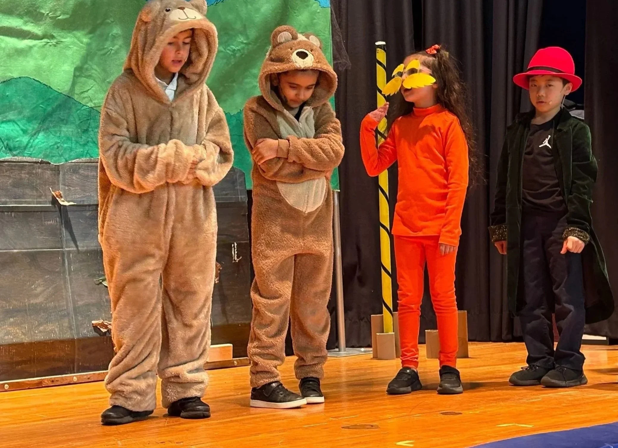 Four children on stage, three dressed in bear costumes with hoods and one in an orange costume with a yellow leaf mask, standing in front of a green and black theatrical backdrop.