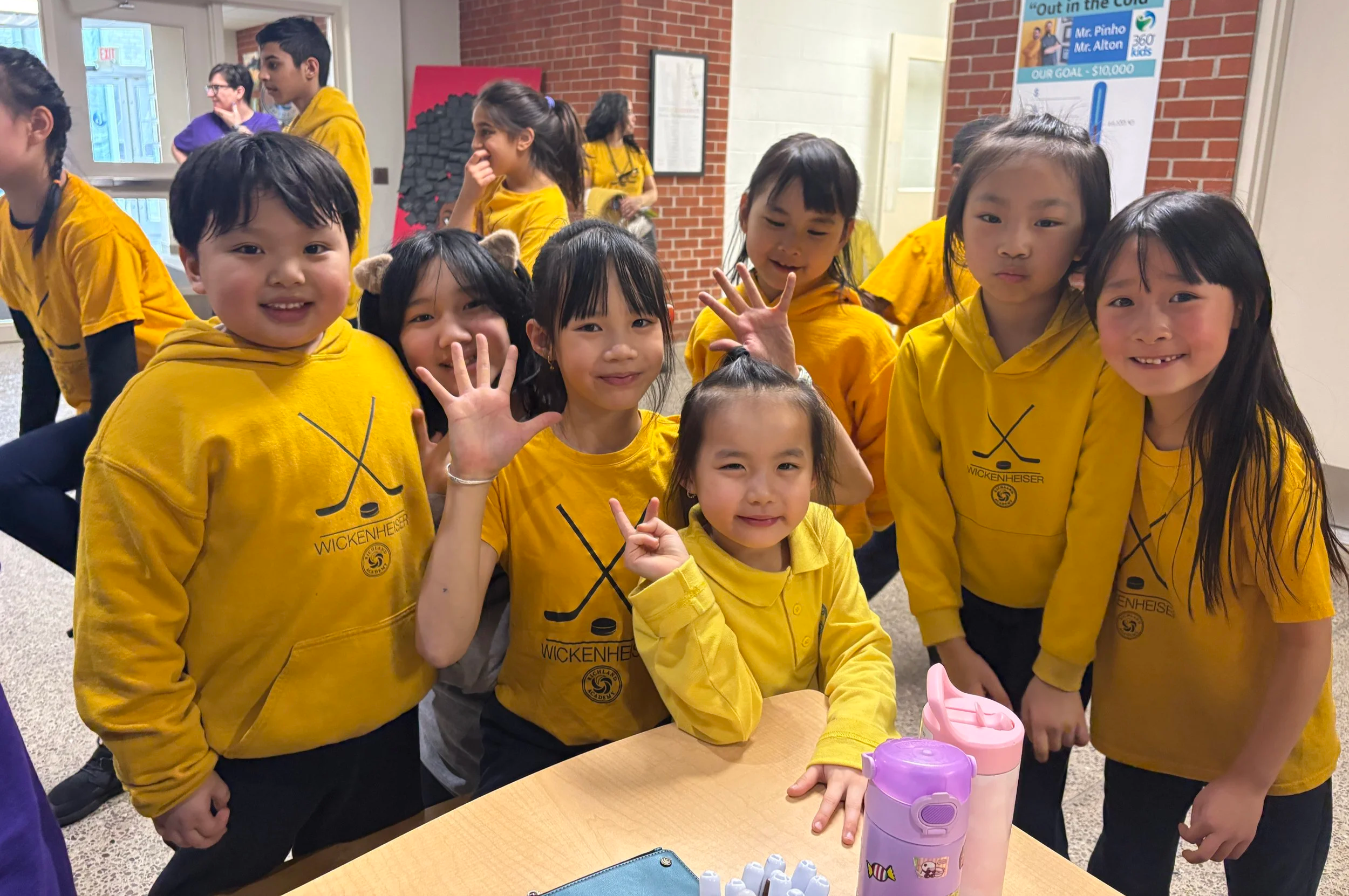 Group of children in yellow hoodies gathered around a table, smiling and posing for the camera in an indoor setting with brick walls and posters in the background.
