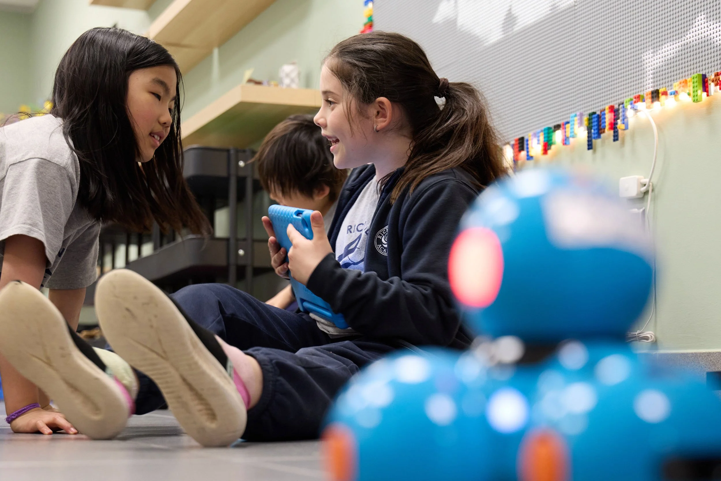 Two girls with brown hair, one Asian and one Caucasian, chatting and smiling while sitting on the floor in a classroom. A third person is partially visible behind them. There is a LEGO wall decoration on the wall behind them and colorful objects on the desk nearby.