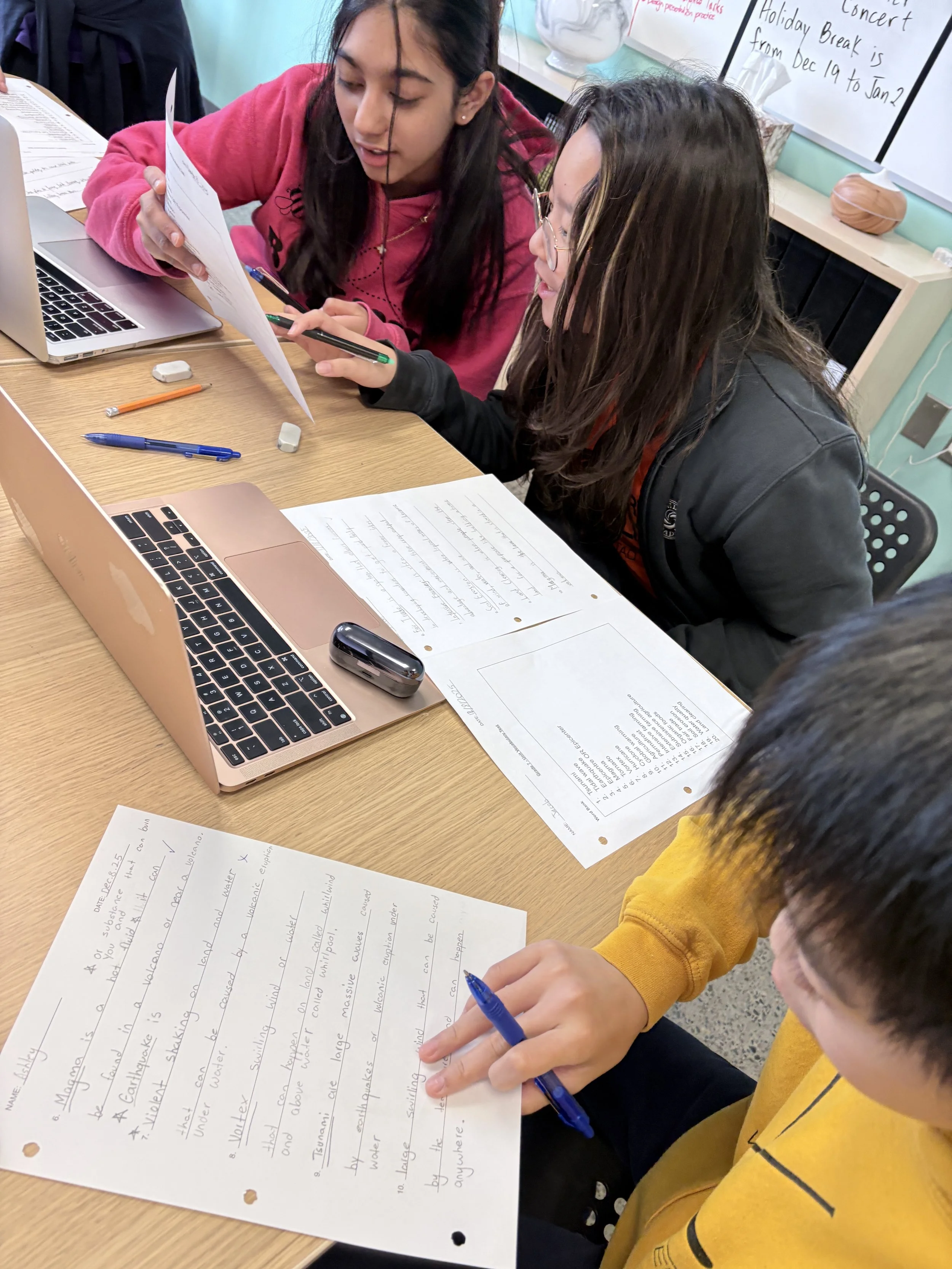 Two students sitting at a desk studying together with laptops, papers, and writing utensils in a classroom setting.