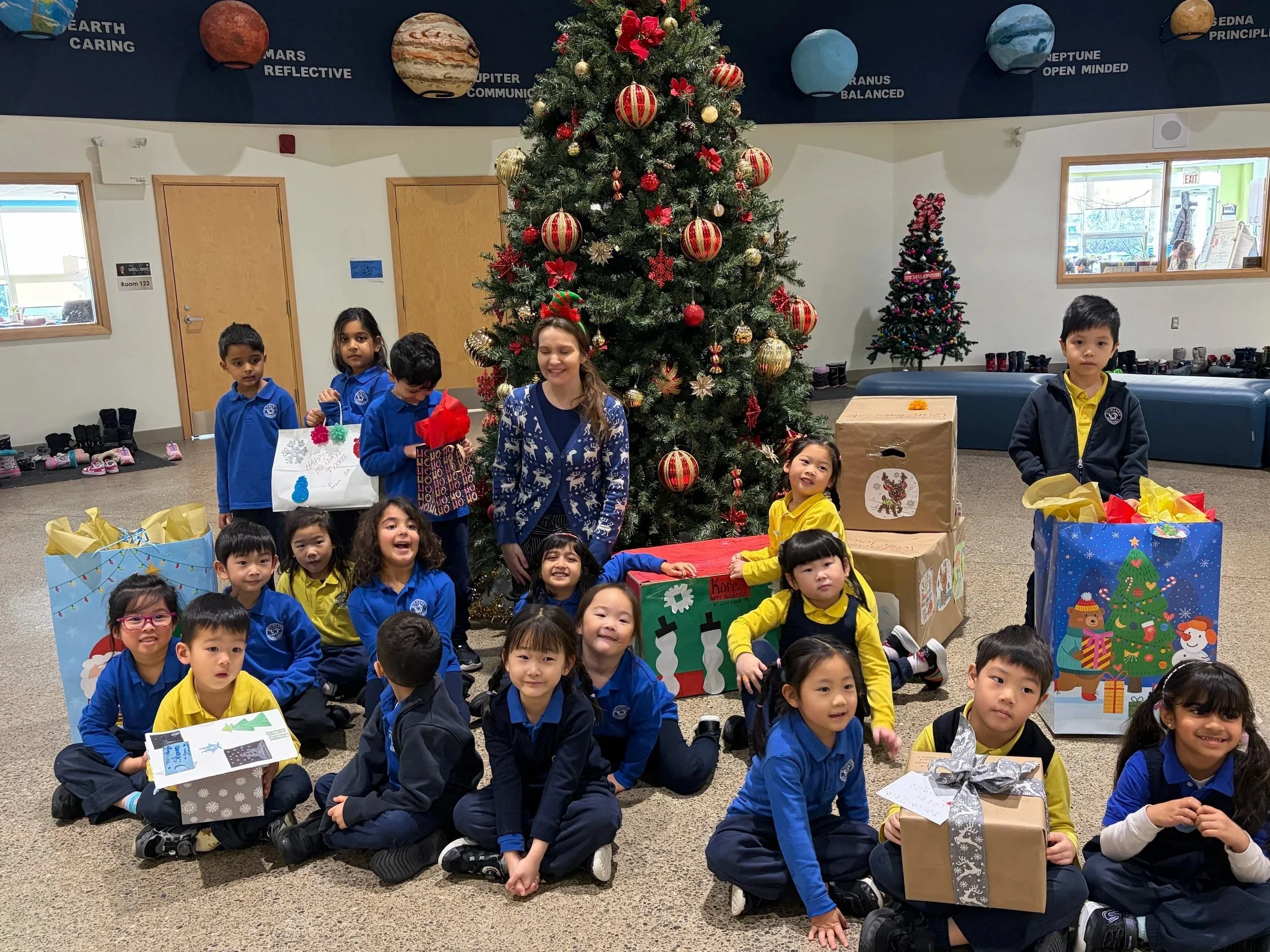 Group of children and a woman gathered around a decorated Christmas tree with presents during a holiday celebration, on a room with space themed decorations on the wall.
