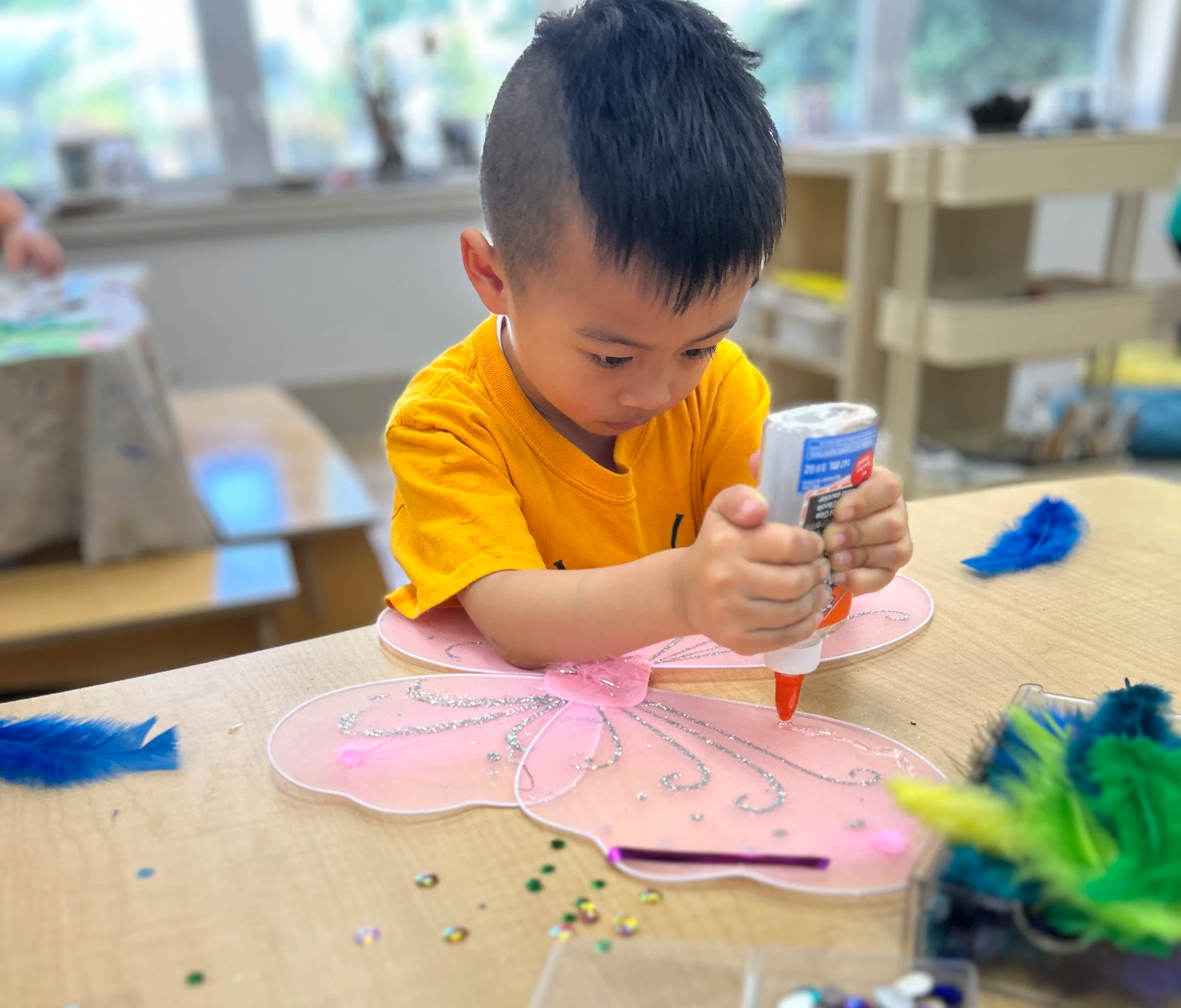 A young boy with short black hair, wearing a yellow shirt, is focused on a craft project. He is sitting at a table decorated with pink and white cutouts and colorful glitter. The background shows a bright room with large windows and shelves.