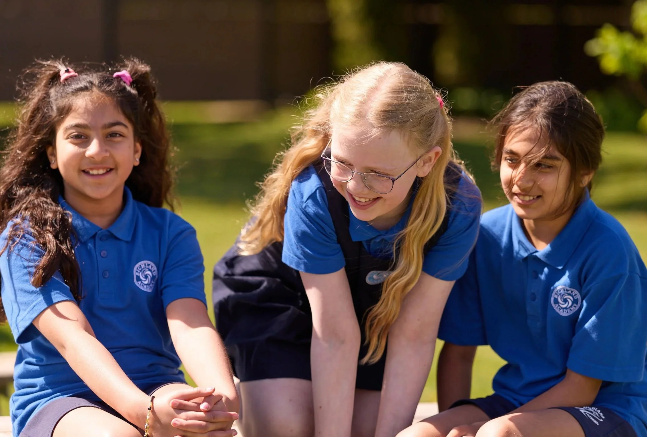 Three young girls wearing blue school uniforms, sitting outdoors and smiling.