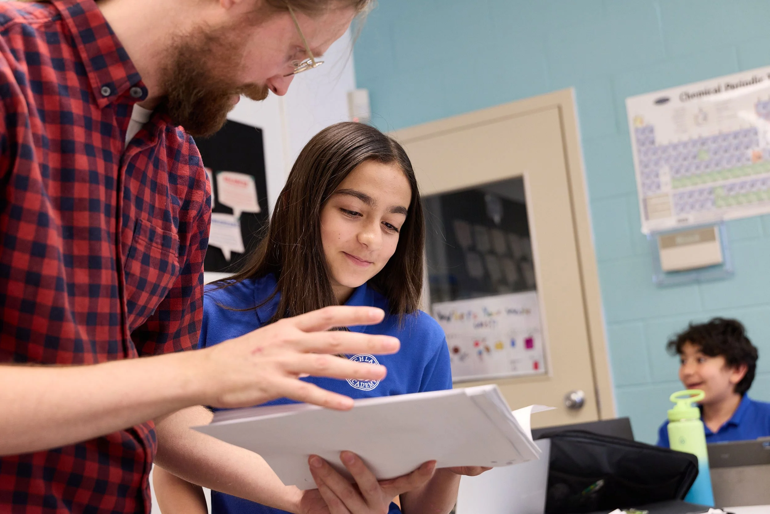 A teacher in a red plaid shirt showing something on a tablet to a girl in a blue shirt in a classroom, with other students in the background.