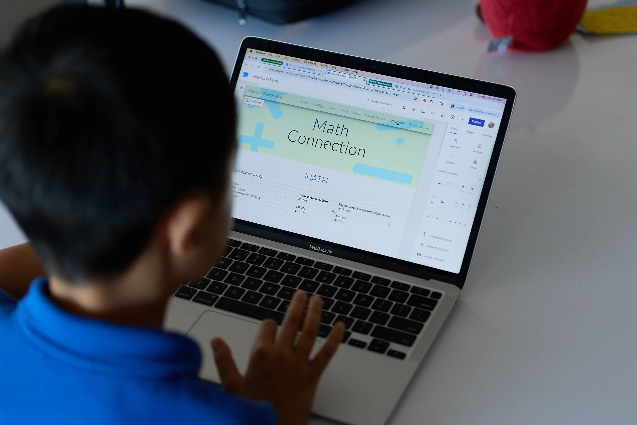 A person wearing a blue shirt using a MacBook Air laptop on a white desk. The screen displays a webpage titled 'Math Connection' with some content and a navigation menu.