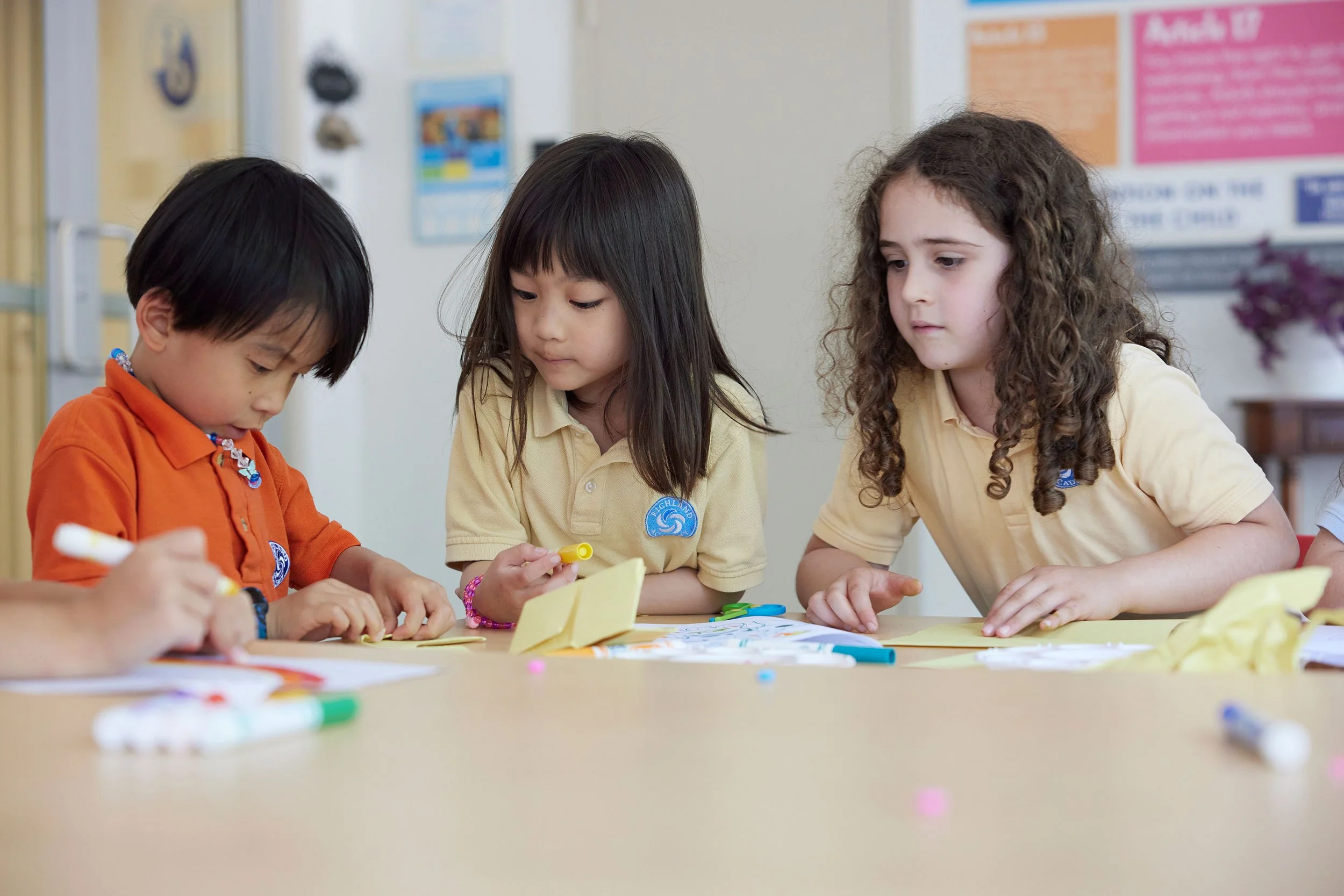 Three young children with light to medium skin tones sitting at a table, engaging in an arts and crafts activity. They are focused on their work, with colorful markers and yellow paper in front of them. The background shows classroom posters and a classroom environment.