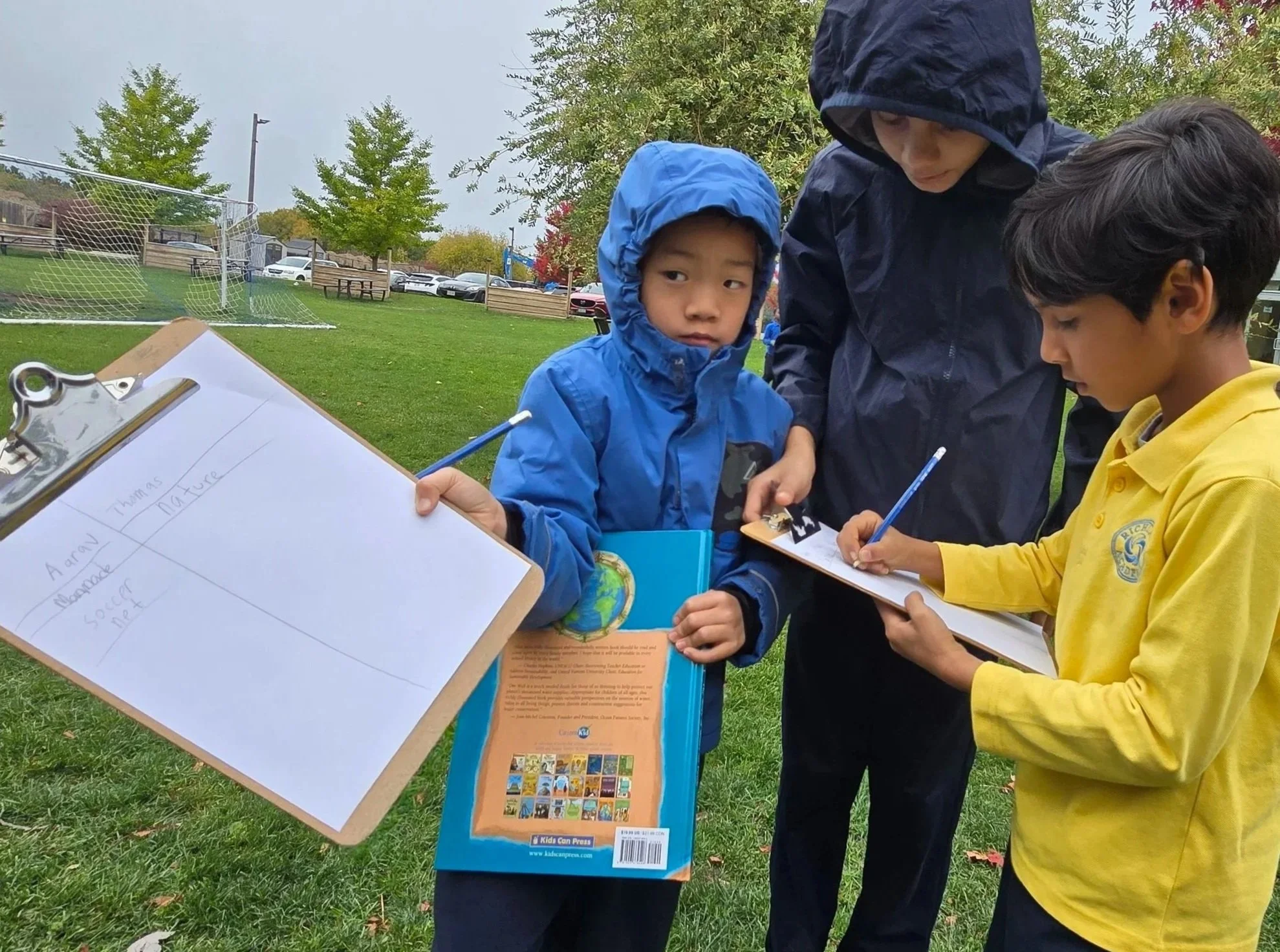 Three boys wearing rain jackets, one in blue, one in dark, and one in yellow, are outdoors at a park on a cloudy day, gathered around a display with clipboards and papers for an educational activity.