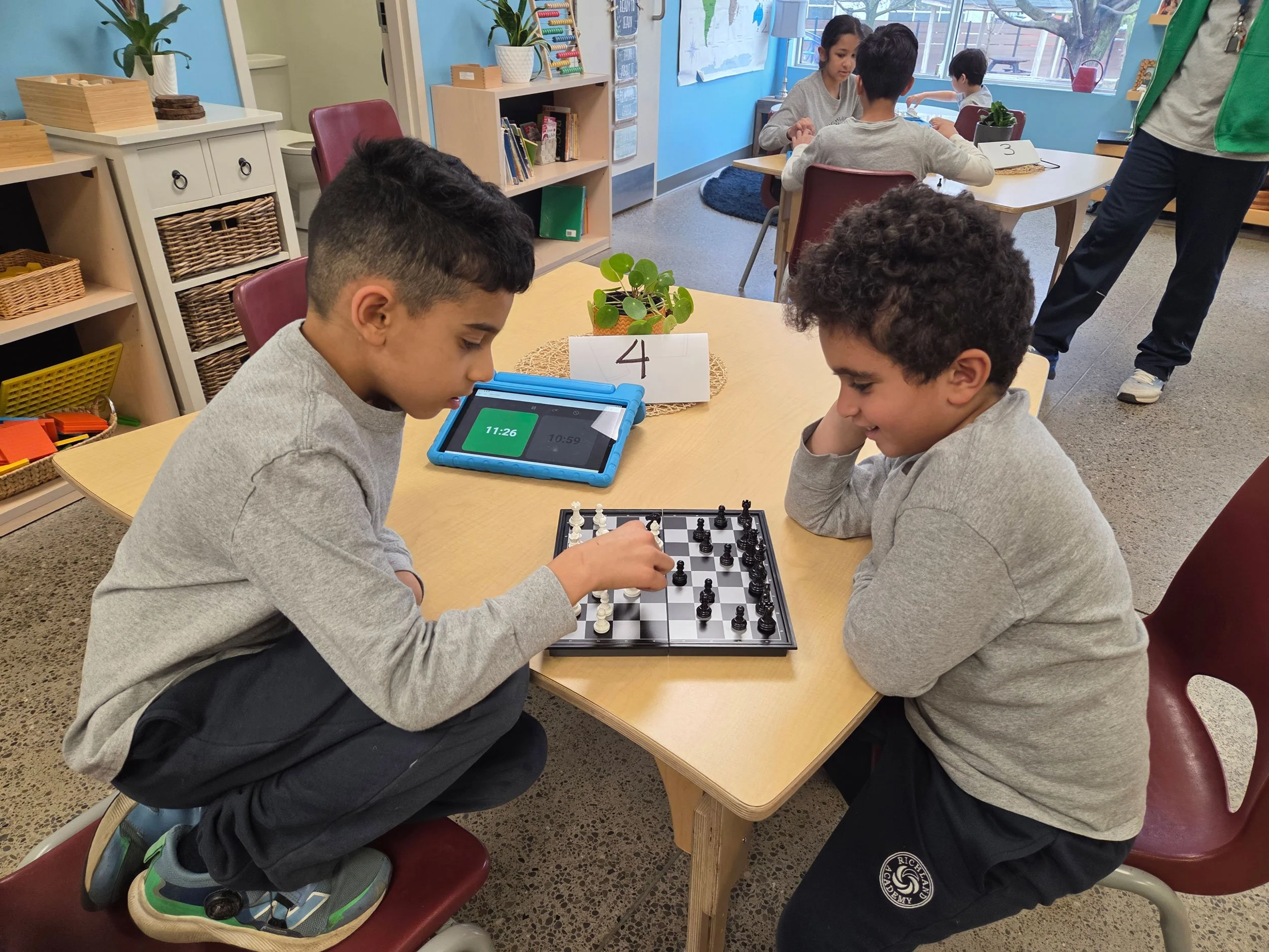 Two boys play chess at a school table while a girl nearby looks on. Other children are seated at tables inside a classroom with blue walls and large windows, with shelves and plants in the background.