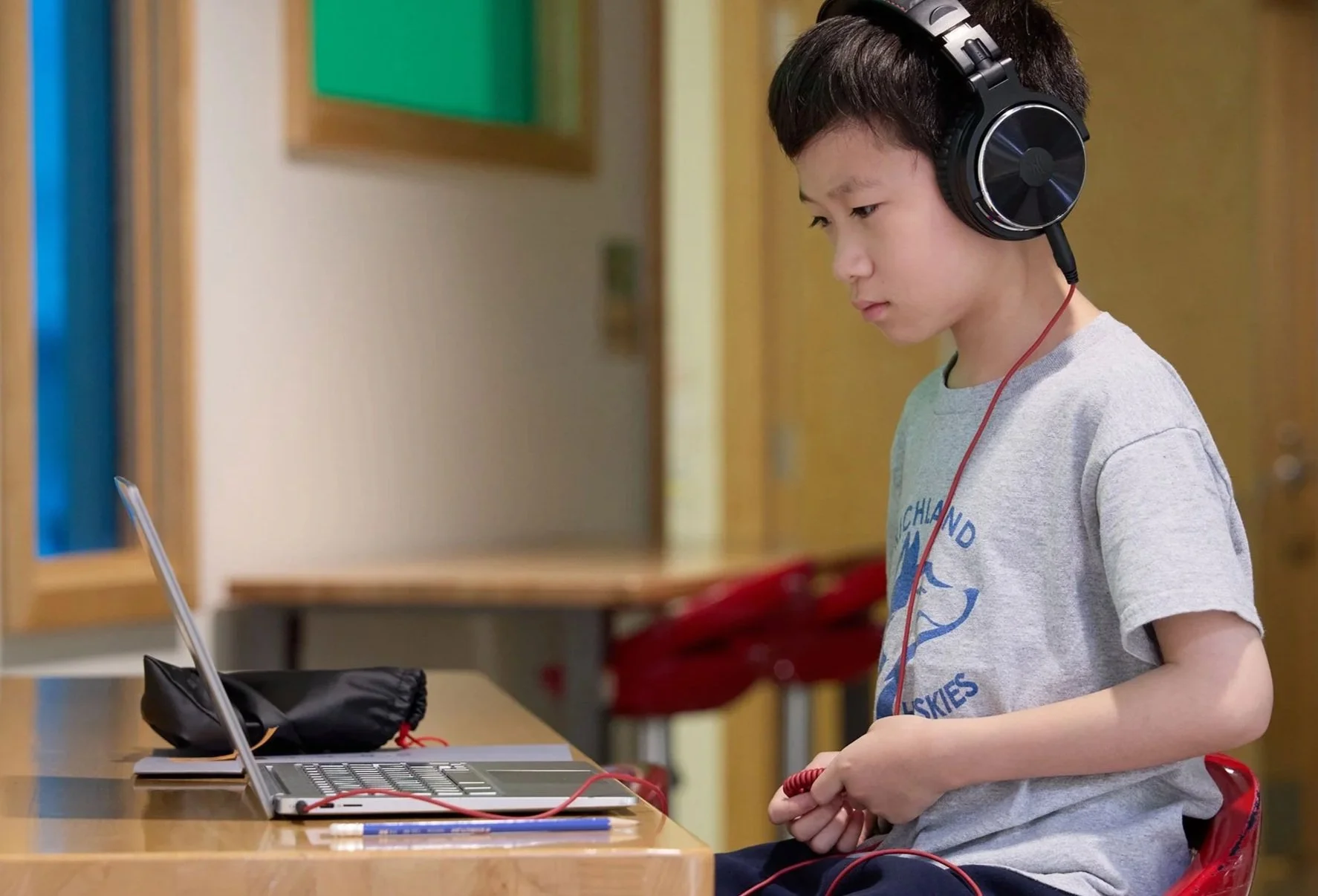 A young boy with dark hair wearing a grey t-shirt and large black headphones sitting at a wooden table in front of a laptop, holding the cable of the headphones.