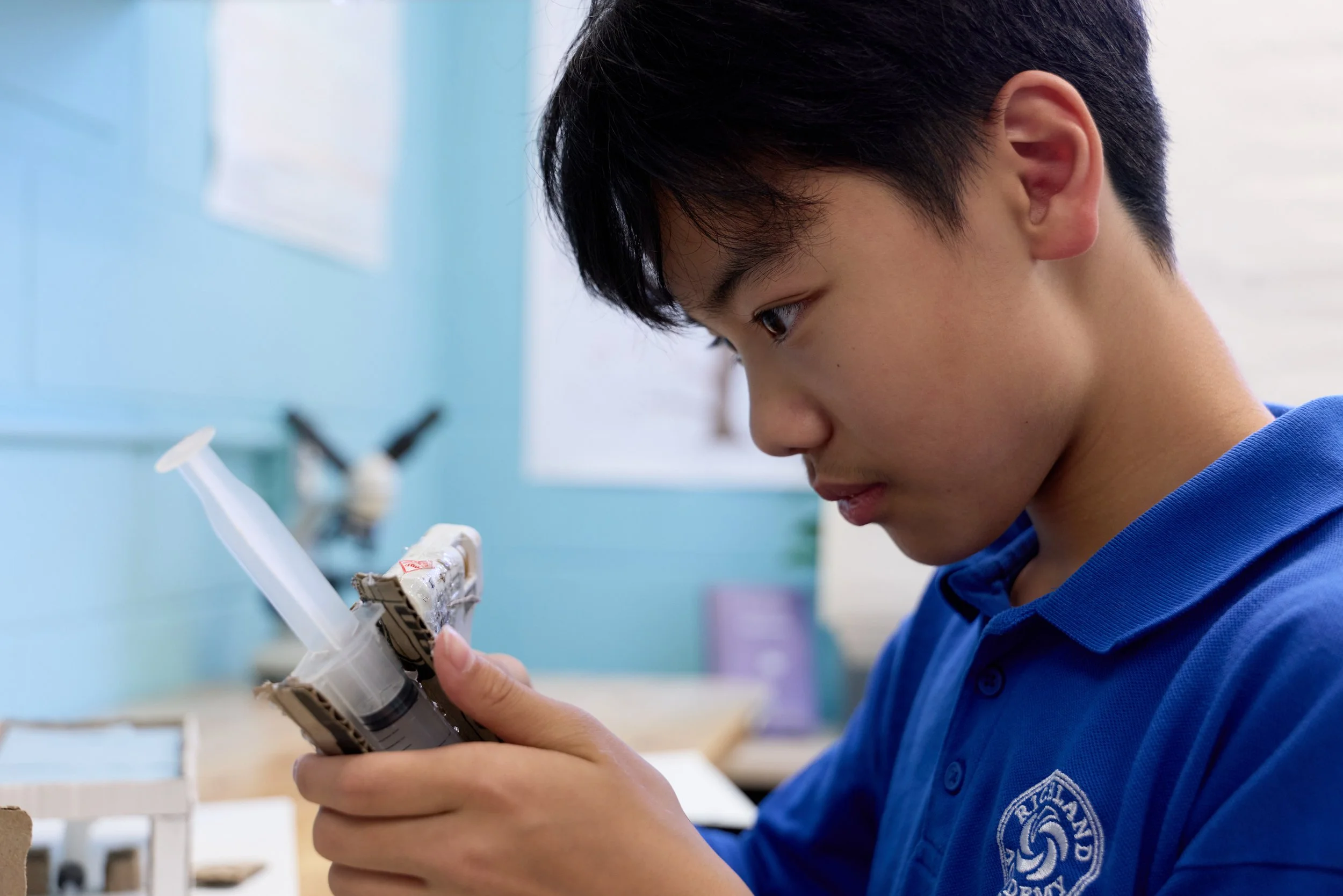 A young person with dark hair, wearing a blue shirt, examining a small device or object closely in a classroom or workshop setting with blue walls and various supplies in the background.