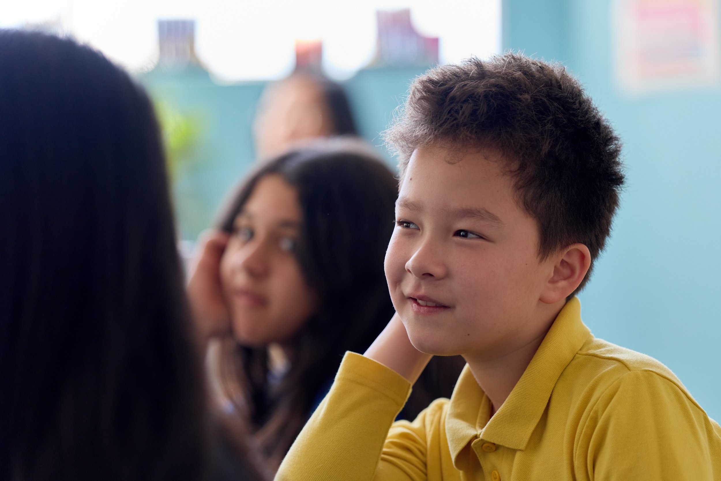 A young boy with curly hair in a yellow shirt is smiling and looking at a person with long dark hair. Two other children are partially visible in the background, one with glasses, in a bright classroom or indoor setting.