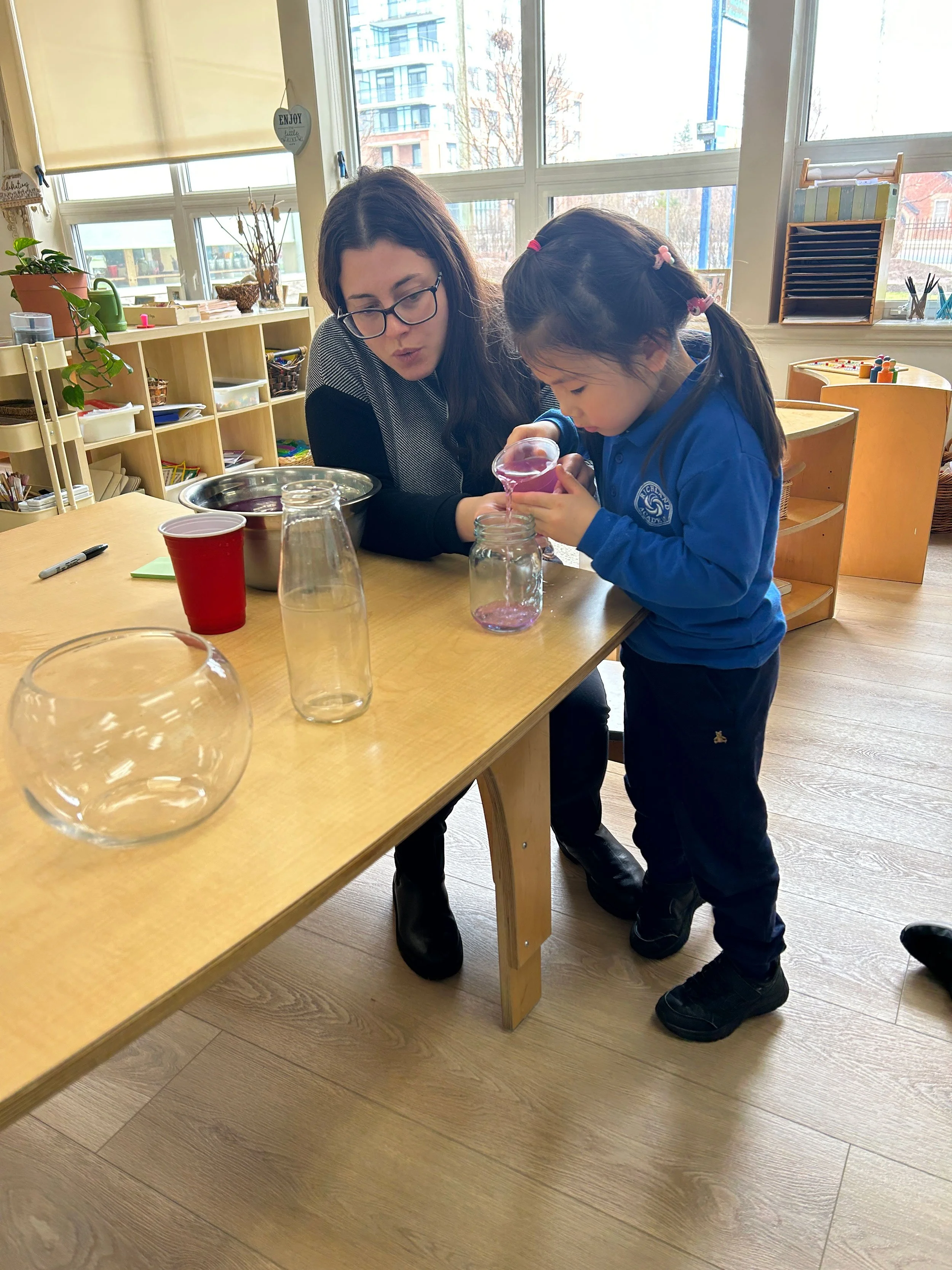 A woman and a girl are pouring liquid from a small funnel into a glass jar on a wooden table. The woman is wearing glasses and a black and white top, while the girl is dressed in a blue school uniform. There are transparent and red cups, and some jars on the table. The background features windows with a view of buildings and trees, and shelves with various supplies.