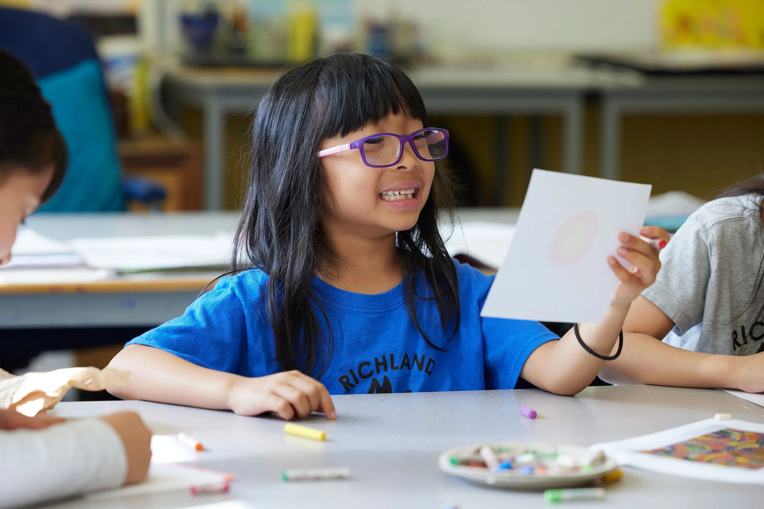 A young girl with long black hair, wearing purple glasses and a blue shirt, is sitting at a desk holding a piece of white paper with a colorful circle drawing, in a classroom setting with other students and school supplies visible.