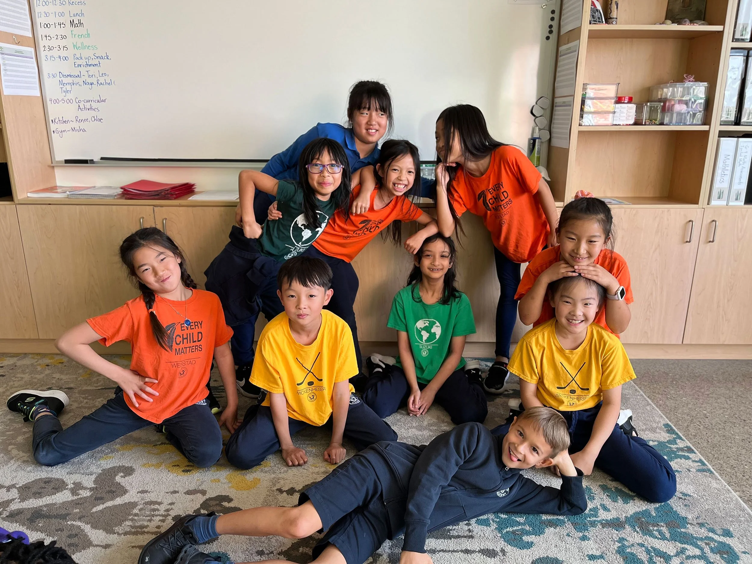 Group of children and a teacher in a classroom, smiling and posing for a photo, with a whiteboard and shelves behind them.