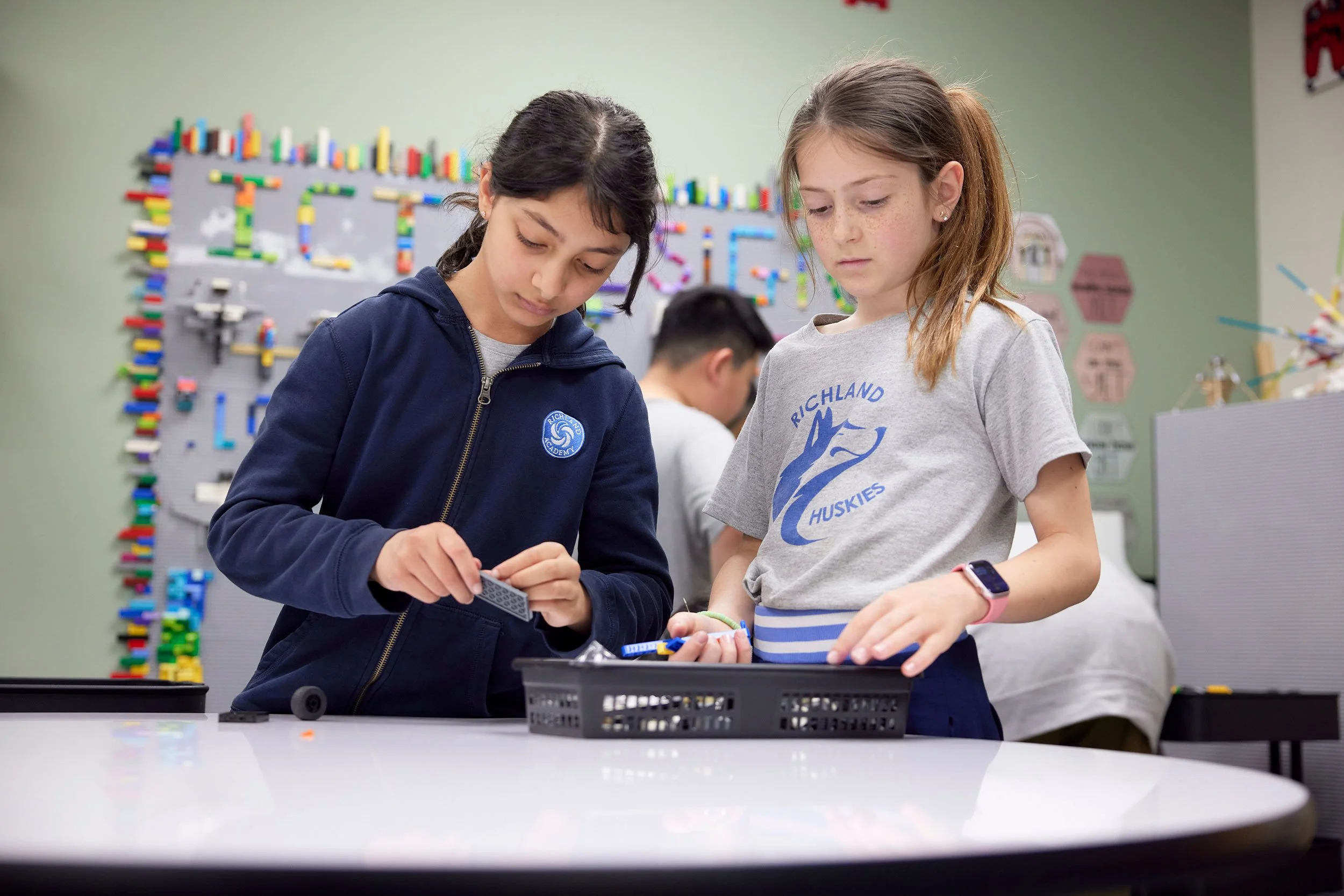 Two girls working on a LEGO project at a table in a classroom. One girl is wearing a dark blue sweatshirt with a school logo, and the other is wearing a gray T-shirt with a blue husky logo. In the background, a colorful LEGO display and a pegboard with LEGO bricks are visible.