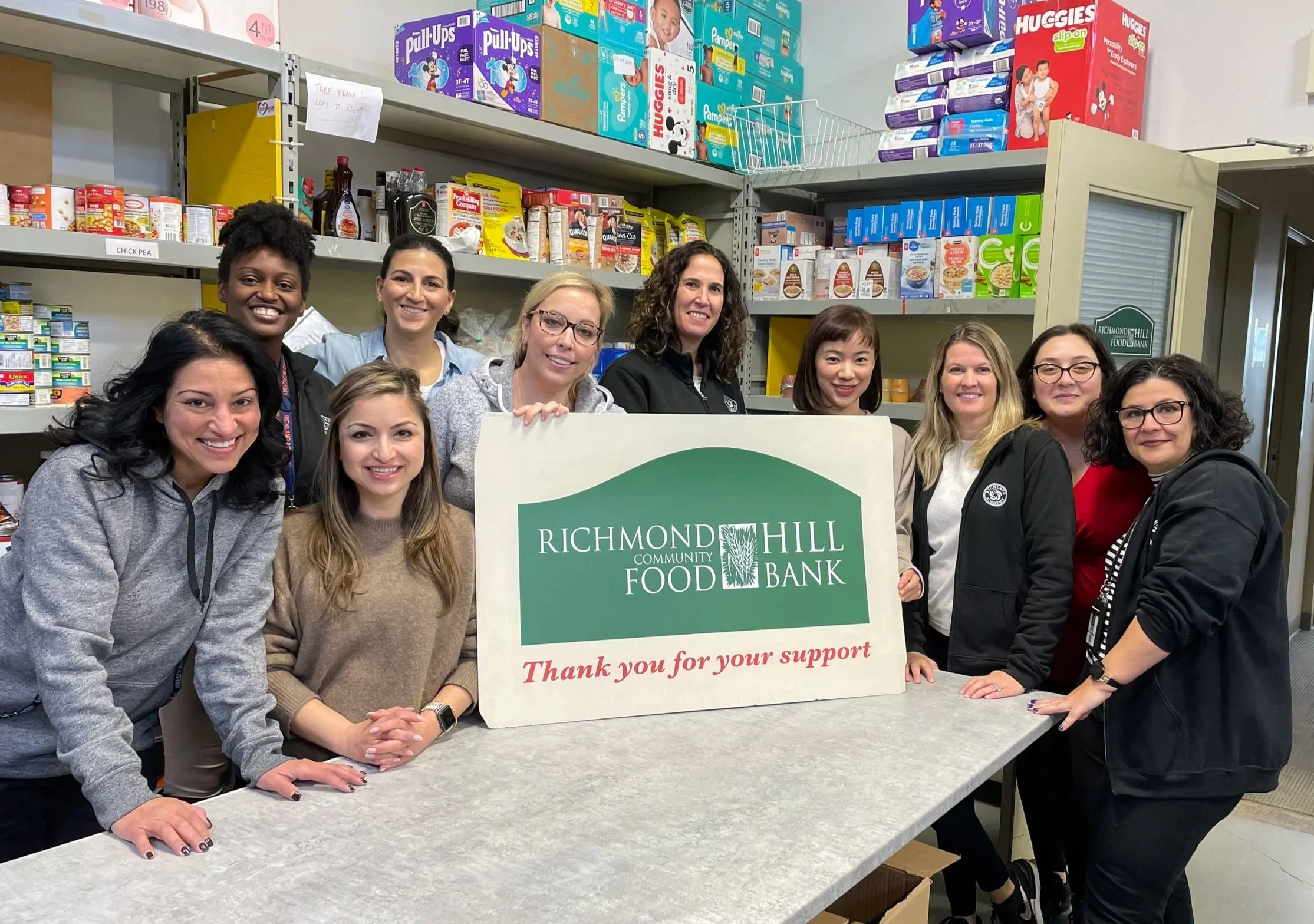 Group of nine women gathered behind a counter in a food bank, smiling, with a Richmond Hill Community Food Bank sign in front, surrounded by shelves of canned goods and boxes of diapers.