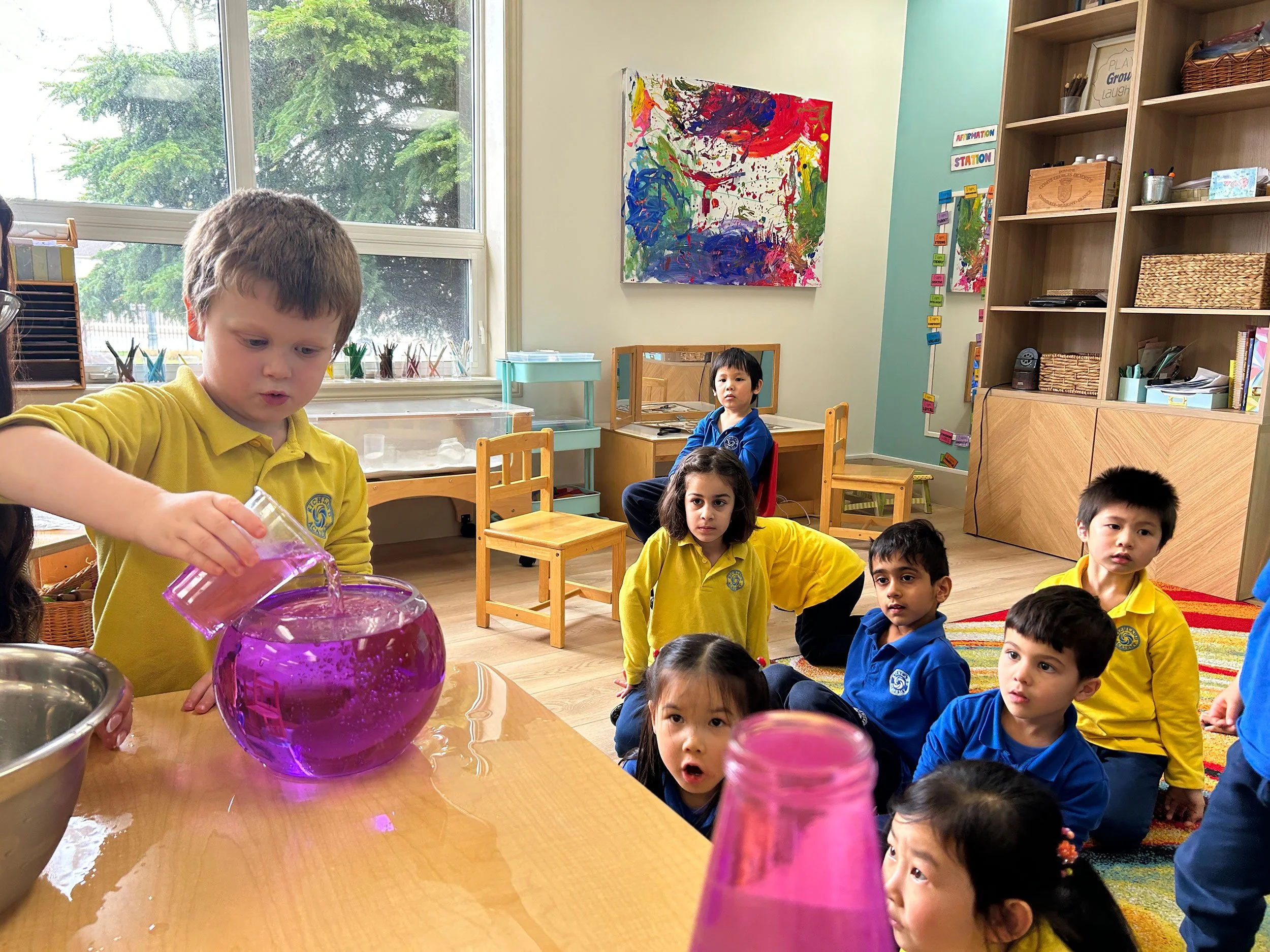 Children in a classroom sitting on the floor while a boy in a yellow uniform pours liquid into a purple sphere-shaped container, with colorful abstract artwork and shelves in the background.
