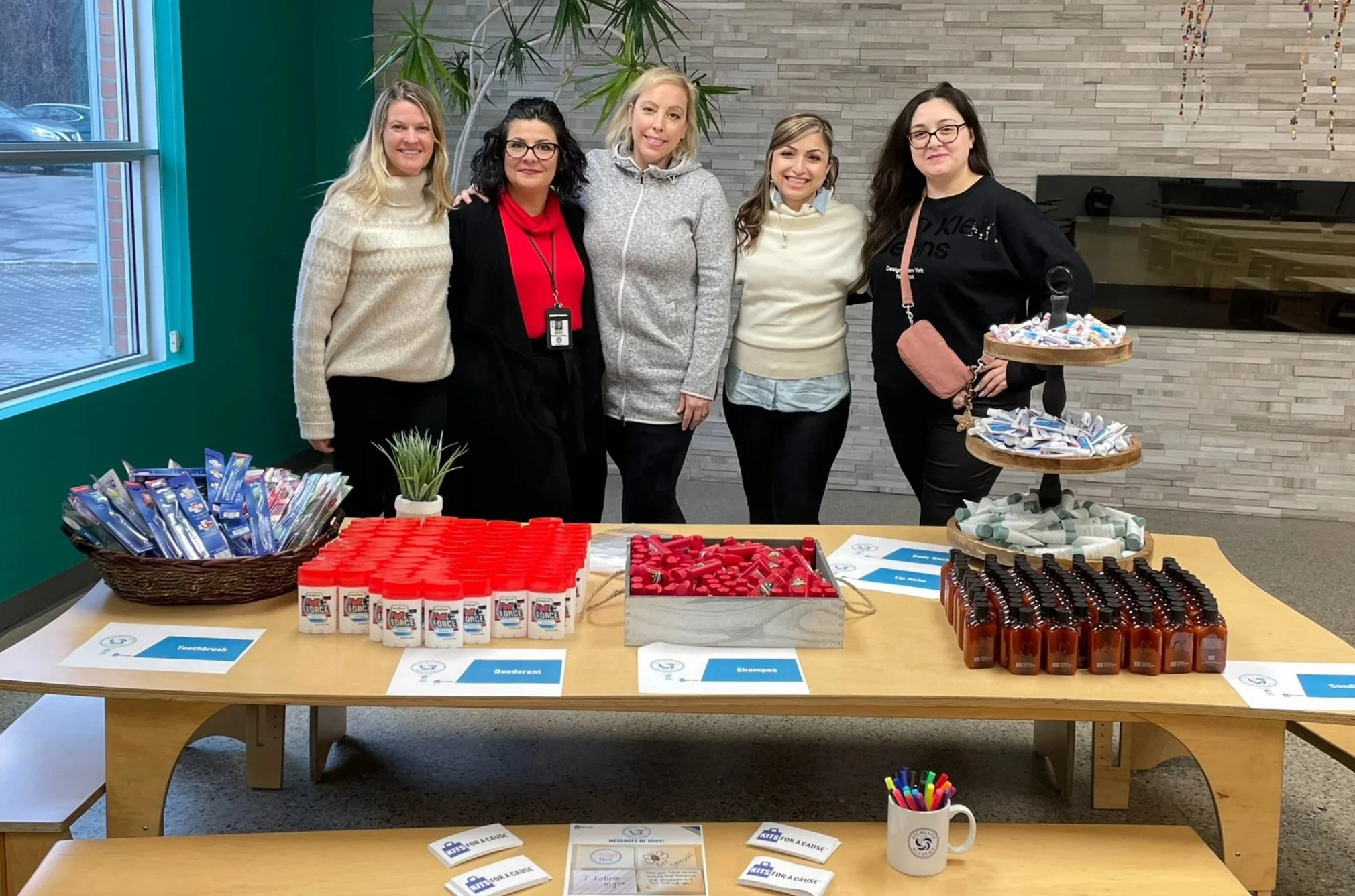 Group of five women standing behind a table with supplies for a fundraising or awareness event, including glue, deodorant, shampoo, and lip balm, in an indoor setting with a gray brick wall and large window.