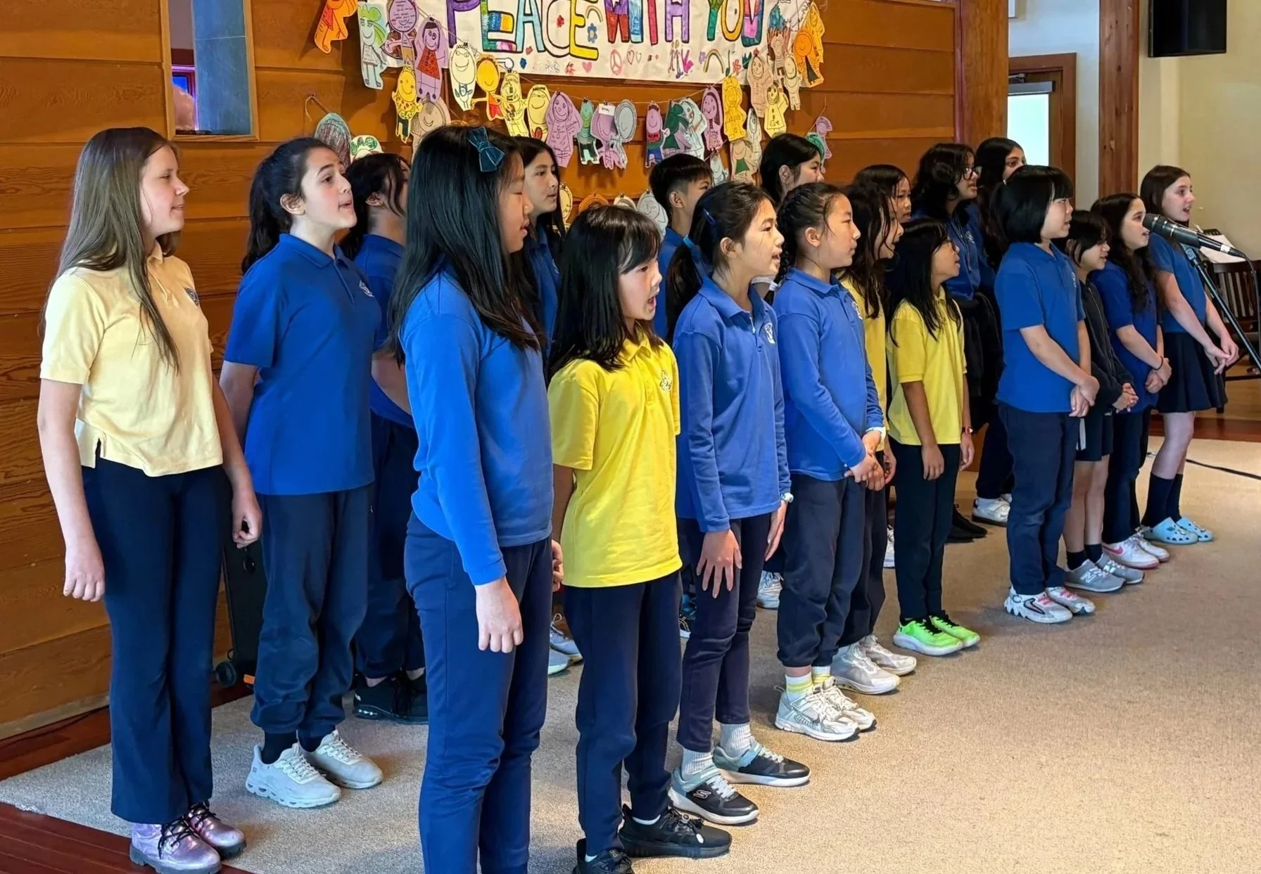 A group of children standing in a line, singing or performing indoors. They are wearing school uniforms in yellow, blue, and black. There is a decorated banner and colorful cutouts of children on the wall behind them.