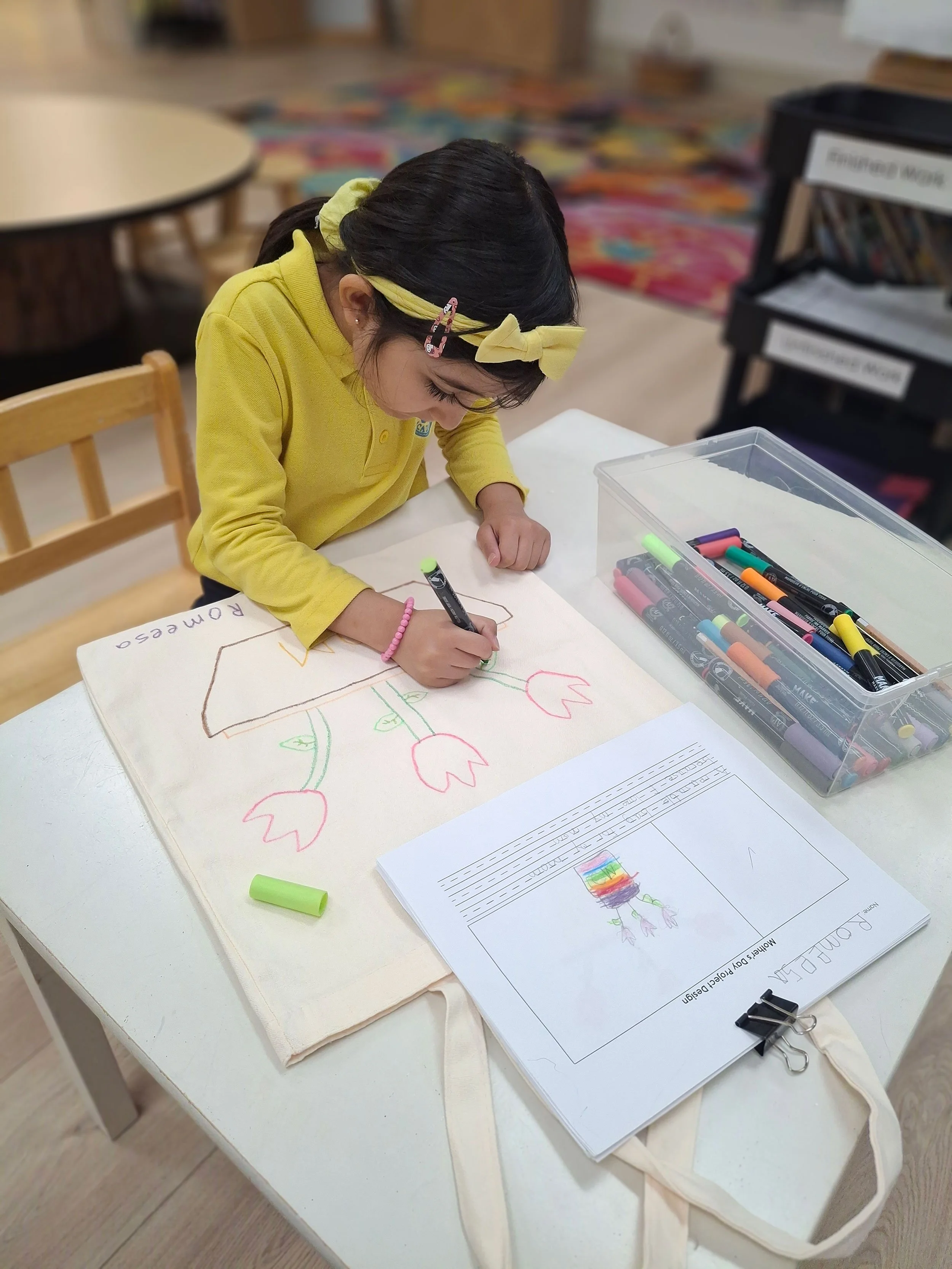 A young girl wearing a yellow headband and yellow shirt drawing a colorful picture of a rainbow, a flower, and the word "romero" on a piece of paper at a white table in a classroom or activity room.