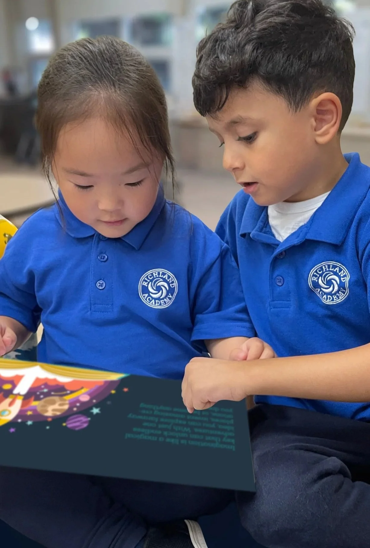 Two young children, a girl and a boy, sitting together and reading a colorful book, both wearing blue shirts with a 'Richland Academy' logo.