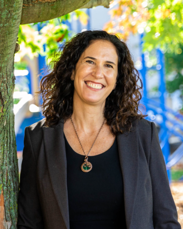 A woman with curly dark hair smiling outdoors, wearing a black top and blazer, standing next to a tree with autumn leaves in the background.