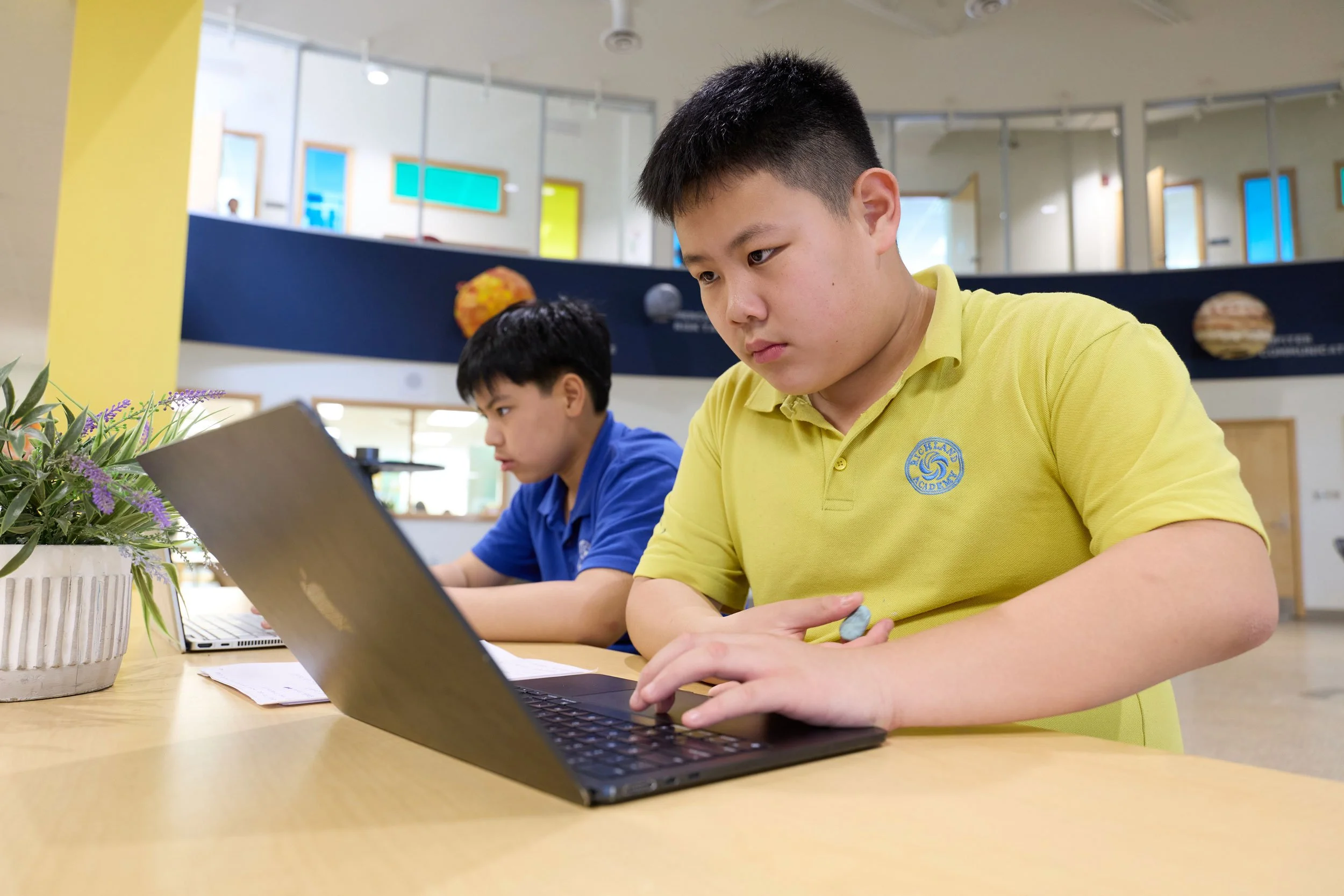 Two boys studying at a table in a bright classroom, one in a yellow shirt and the other in a blue shirt, with a potted plant in the foreground.