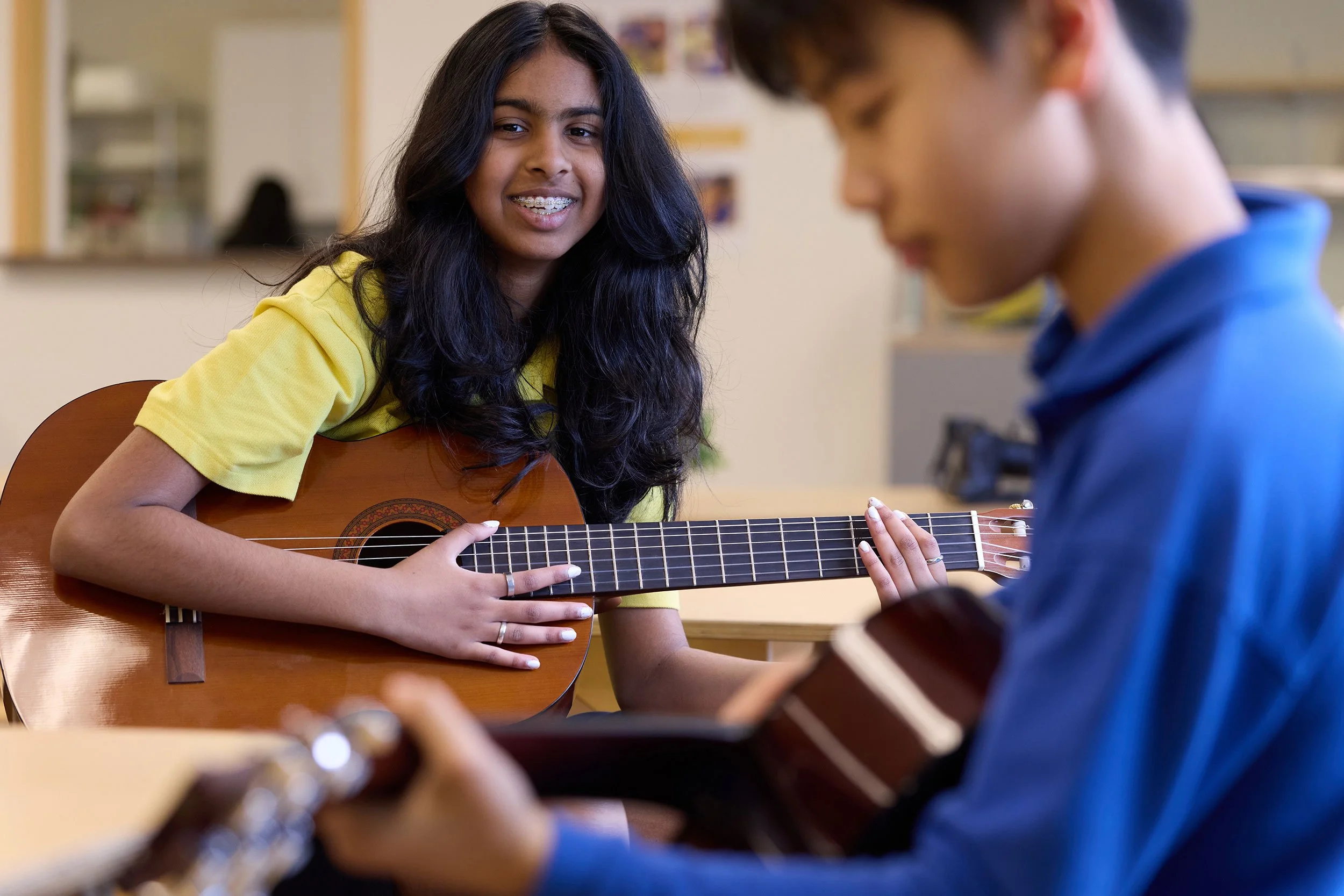 A girl with long dark hair and braces playing an acoustic guitar, sitting at a table with another person whose face is out of focus.