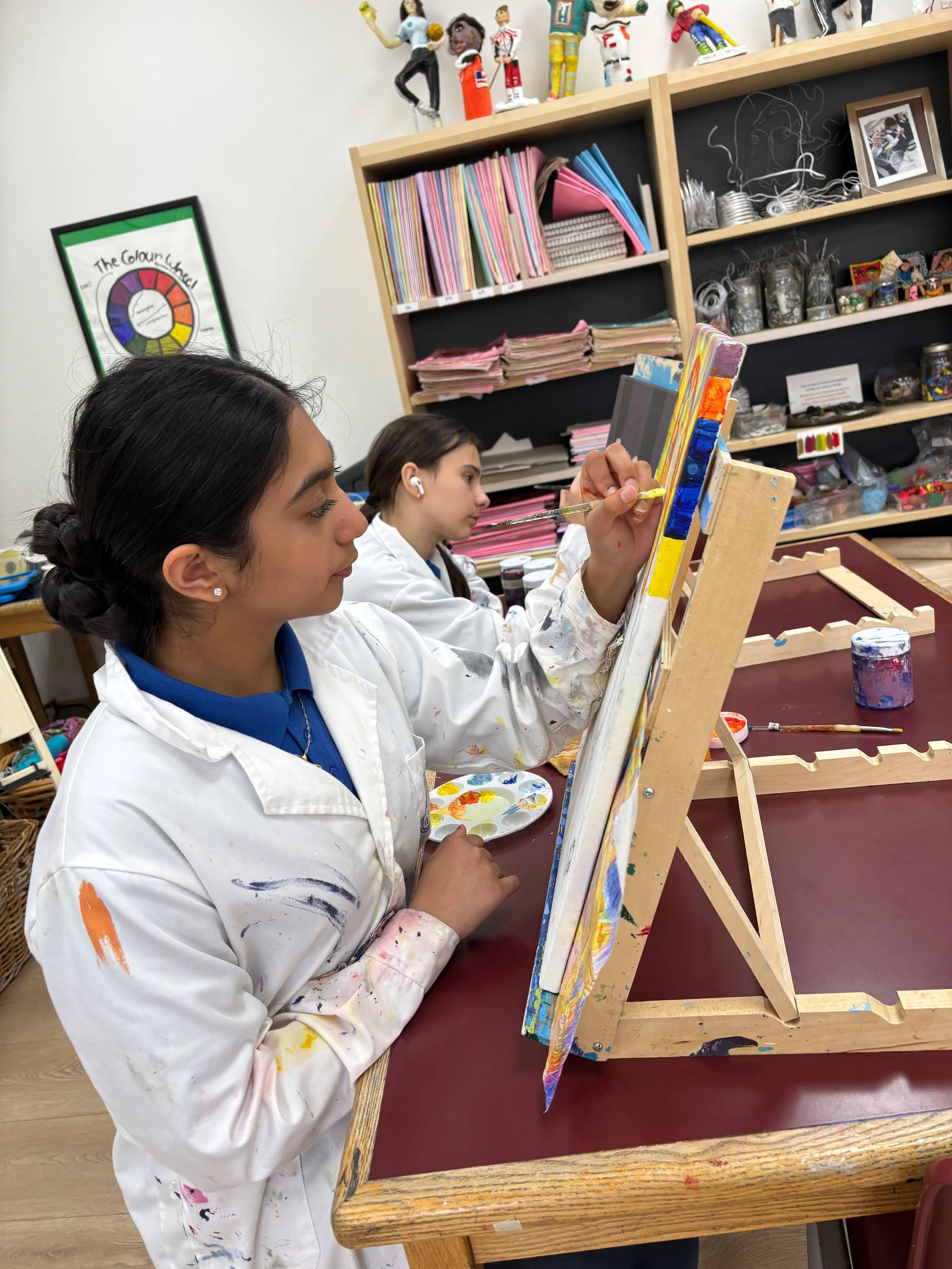 Two young girls painting on canvases set on easels in an art classroom. The girls are wearing white smocks with paint stains. Shelves in the background hold colorful files, paper, and art supplies, and there are sculptures and photographs displayed on top. One girl is holding a paintbrush and palette, focused on her artwork.
