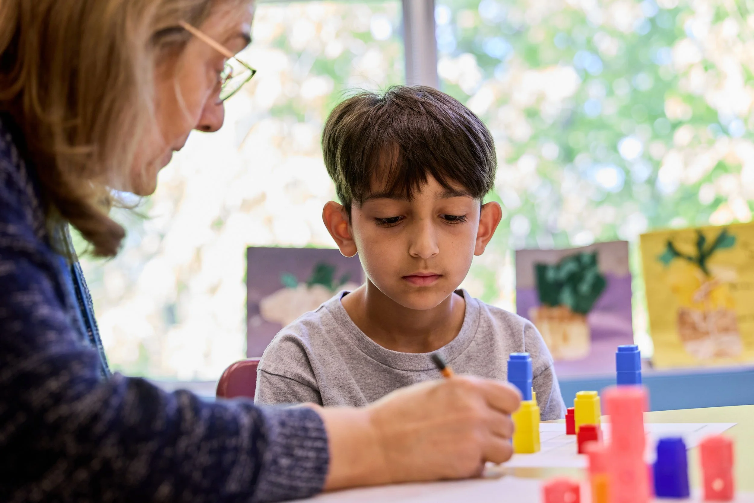 A woman and a young boy playing a board game involving colorful blocks indoors with a sunny window in the background.