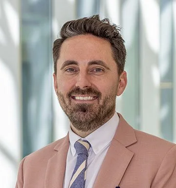 A man with a beard and brown hair smiling, wearing a light brown suit jacket, white shirt, and a blue and white striped tie, standing in a modern office with large windows in the background.