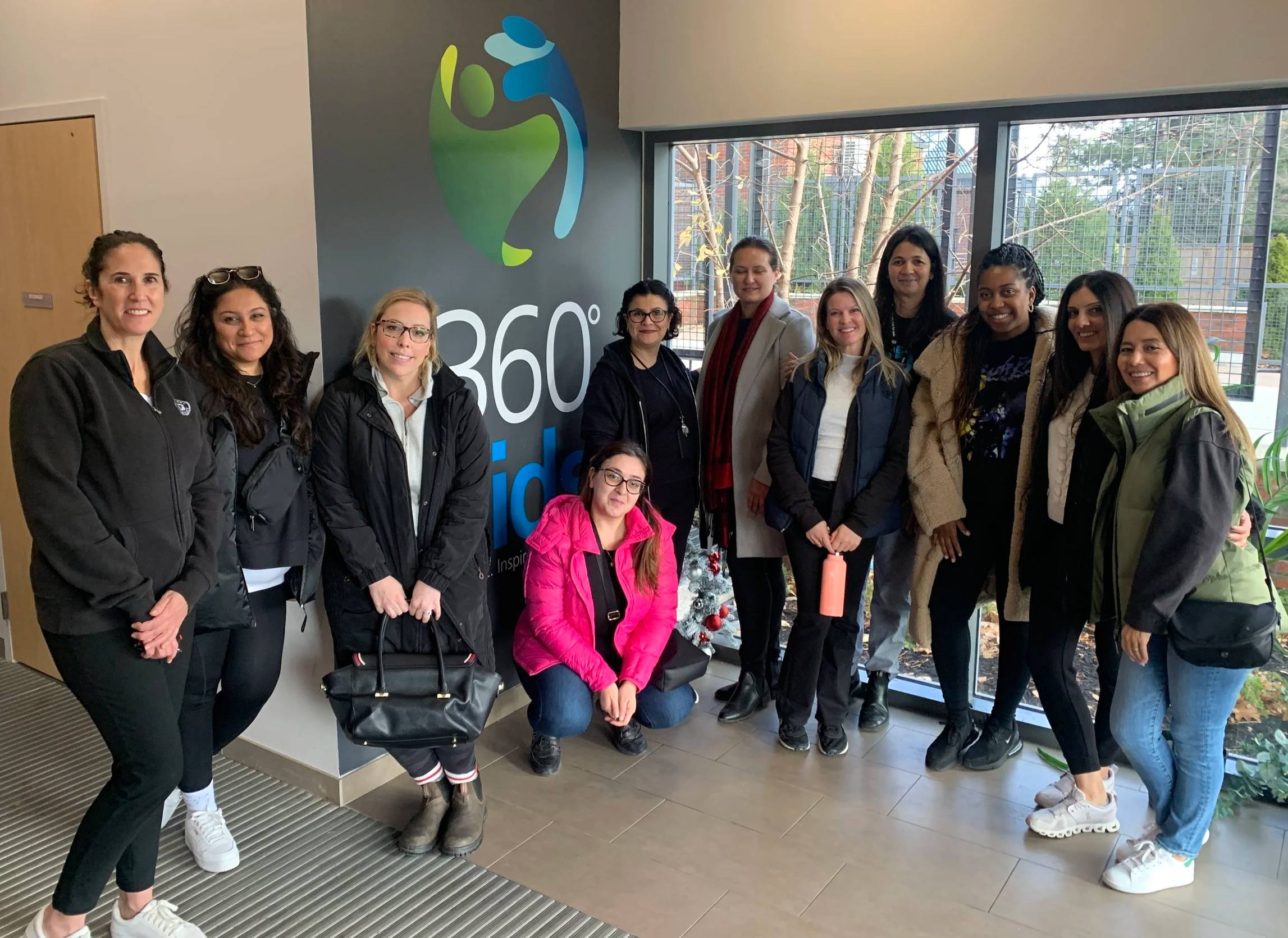Group of women standing indoors near a large window, posing for a photo, with a sign that reads '360°' on the wall behind them.