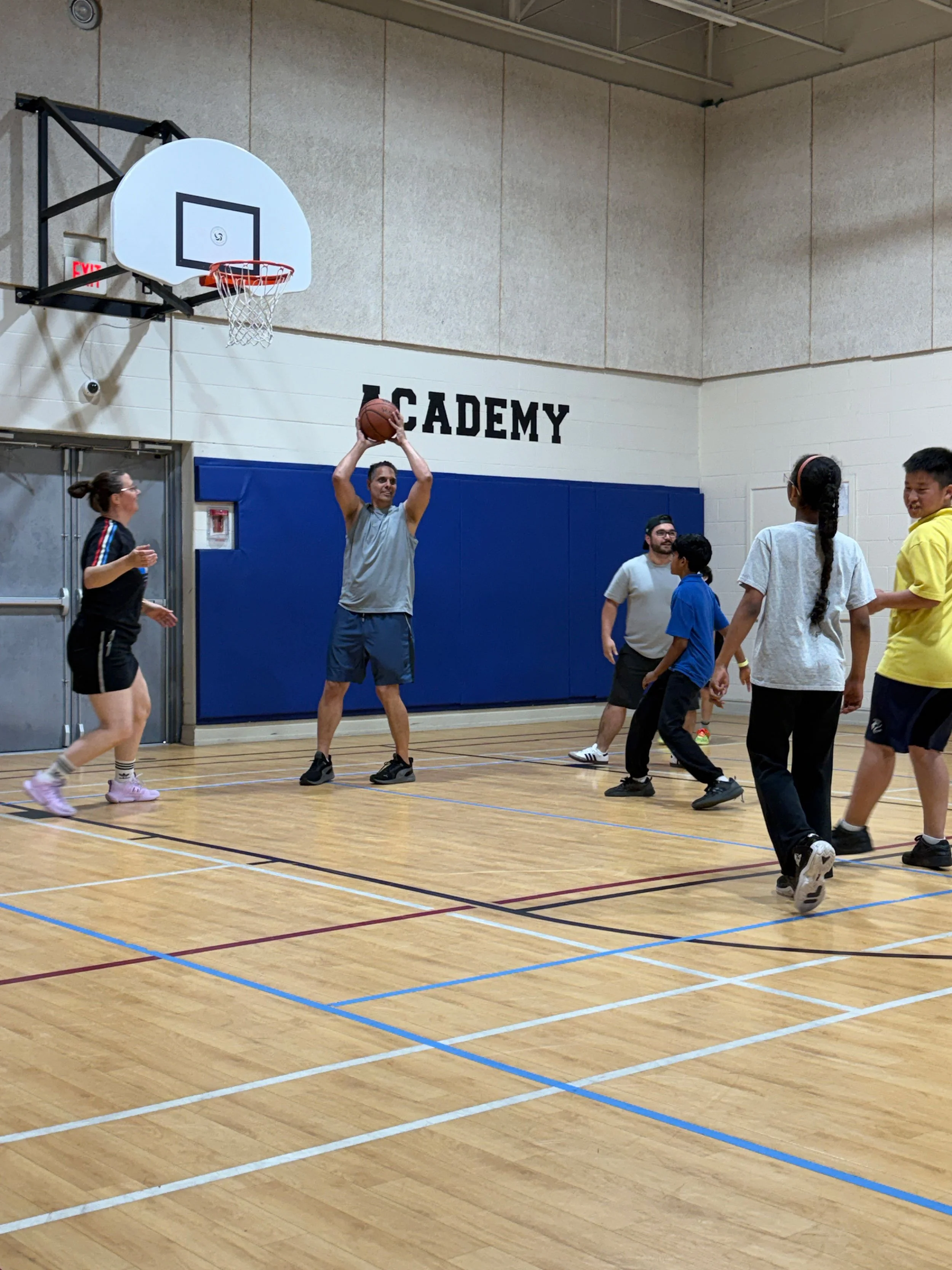 A group of people playing basketball in an indoor gymnasium, with one man shooting a basketball and others watching or moving around.