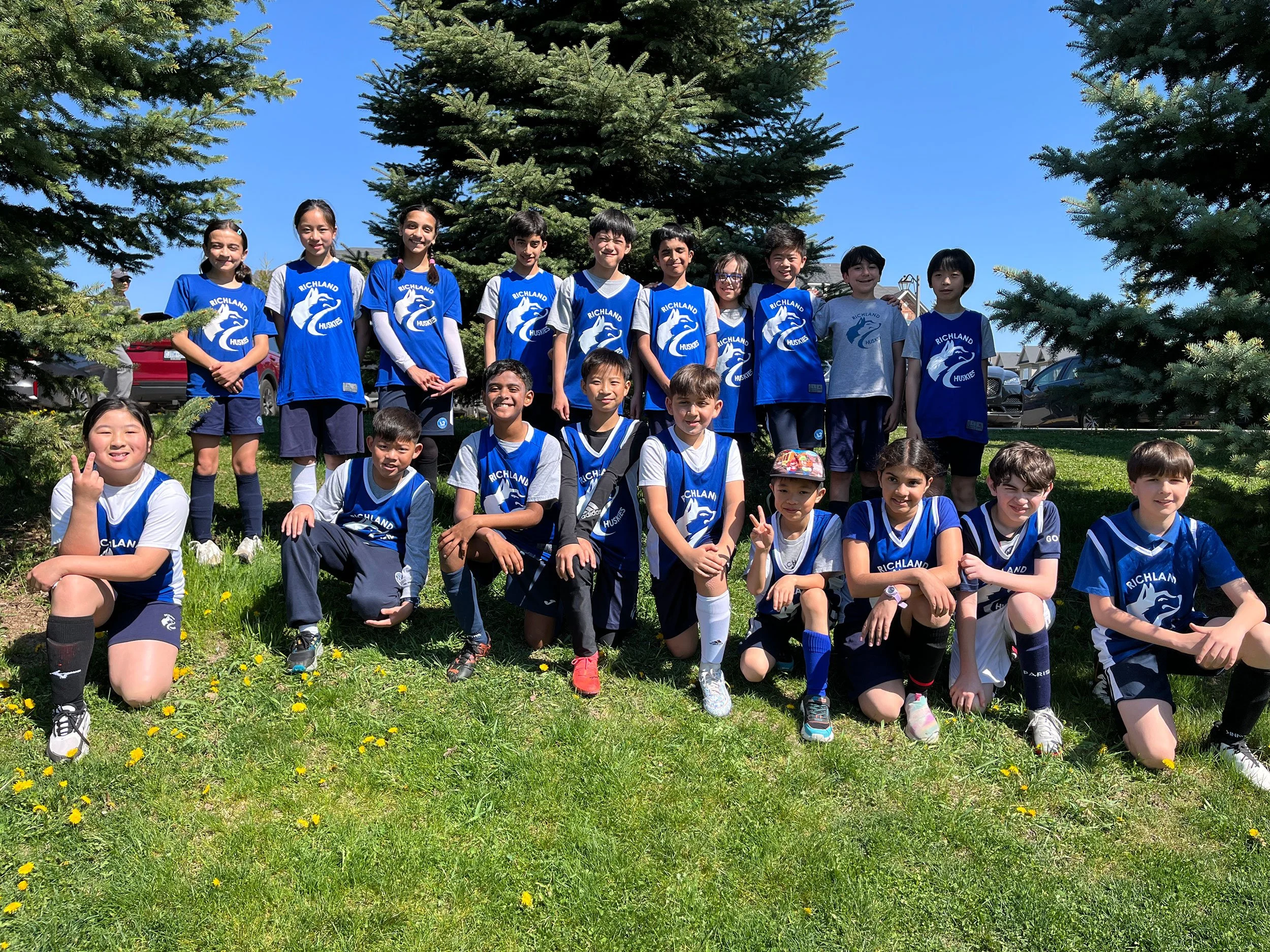 A group of children wearing blue and gray athletic jerseys with 'Richland Huskies' written on them, posing outdoors in front of pine trees on a sunny day. Some children are standing, while others are sitting or kneeling on the grass, smiling and making peace signs.