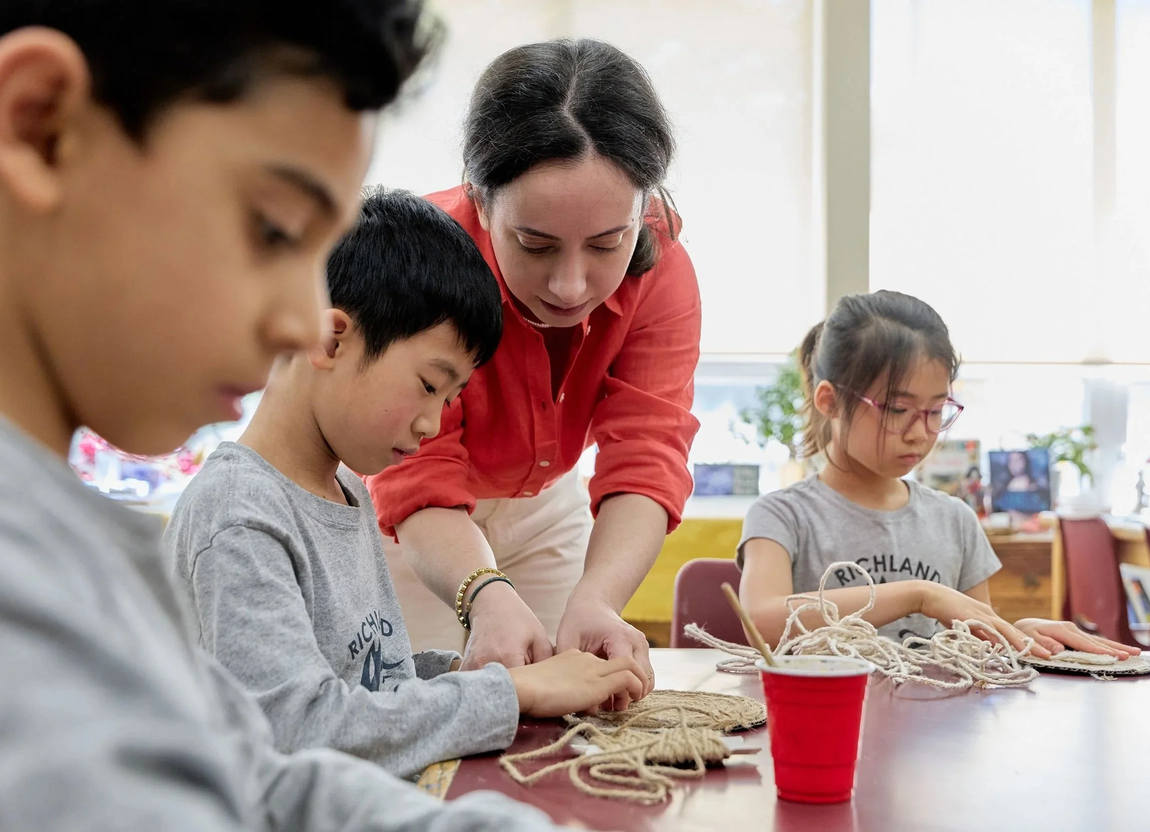 A teacher instructing children in a classroom on a craft project involving yarn. The teacher is leaning over, assisting the children who are focused on their work at a table. The children are wearing casual clothes, and the classroom is well-lit with windows.