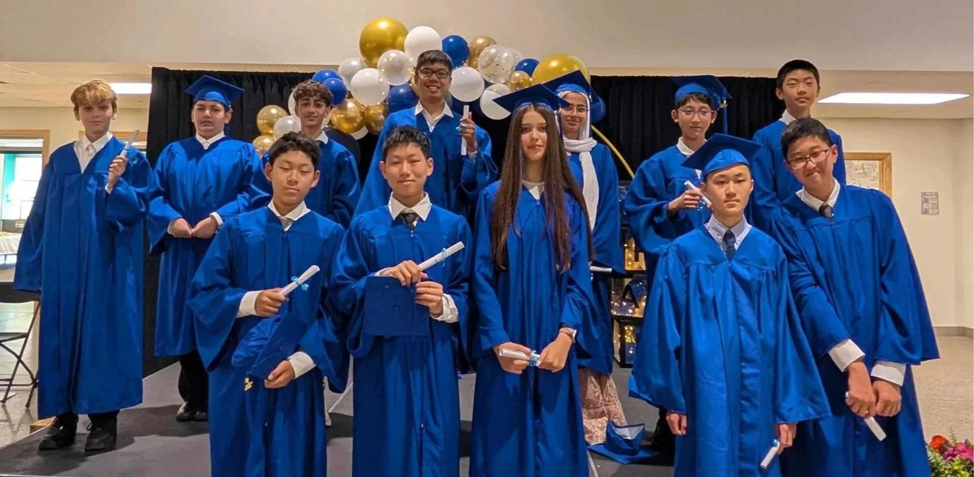 Group of students in blue graduation caps and gowns posing at graduation ceremony, with balloons and decorative backdrop in background.