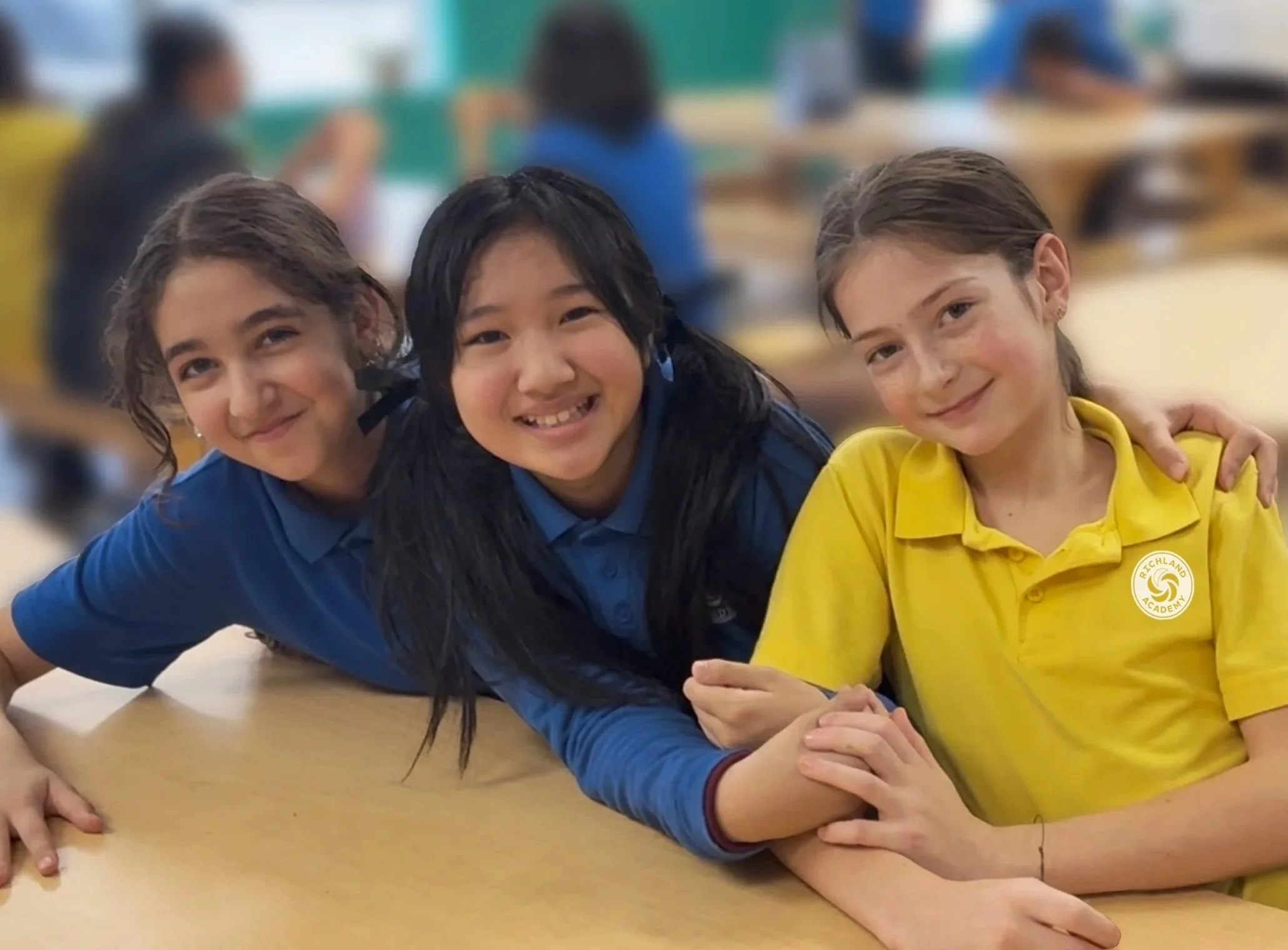 Three girls are sitting at a school desk, arm-in-arm, smiling at the camera in a classroom. The girl in the middle has black hair and is wearing a blue uniform; the girl on the left has brown hair and wears a blue uniform; the girl on the right has light brown hair and is wearing a yellow uniform with a school logo.