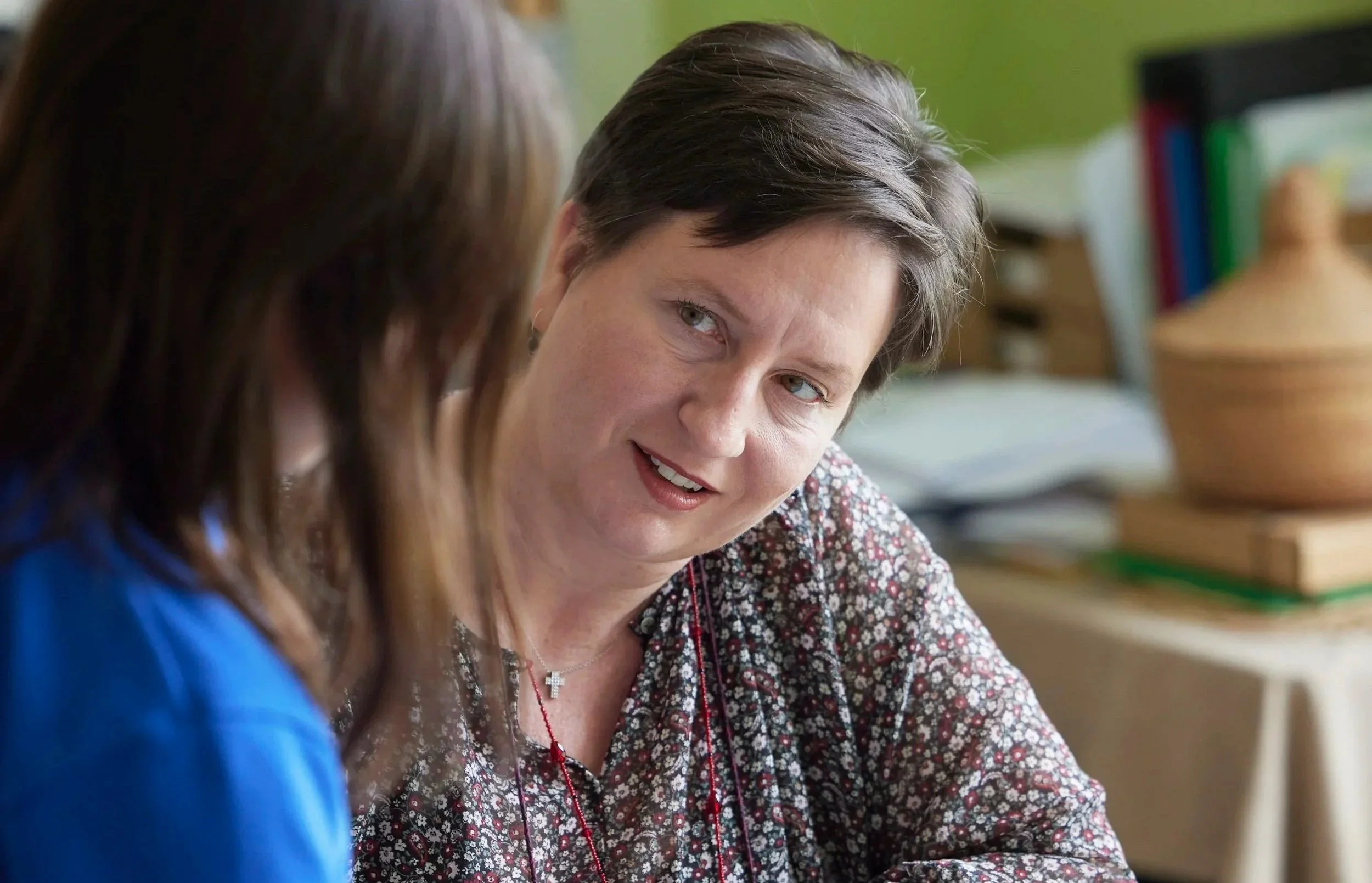 A woman with short brown hair and light skin, wearing a patterned shirt, is looking at a person with long brown hair in the foreground. The scene appears to take place in an office or classroom with shelves and binders in the background.